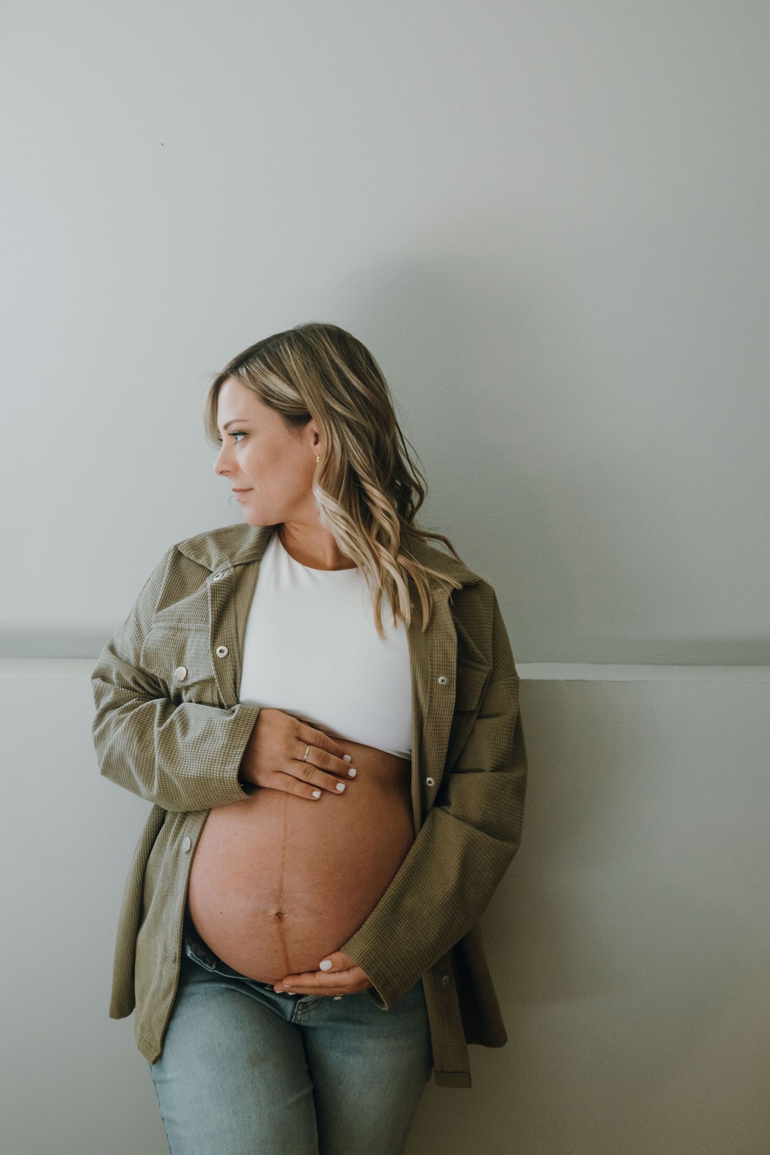 Pregnant woman in olive jacket and jeans cradling baby bump against white wall.