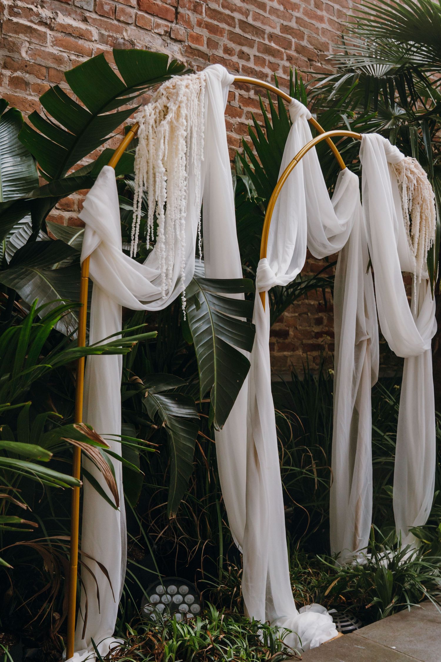 Elegant gold wedding arch draped with white fabric and macrame fringe against tropical greenery.