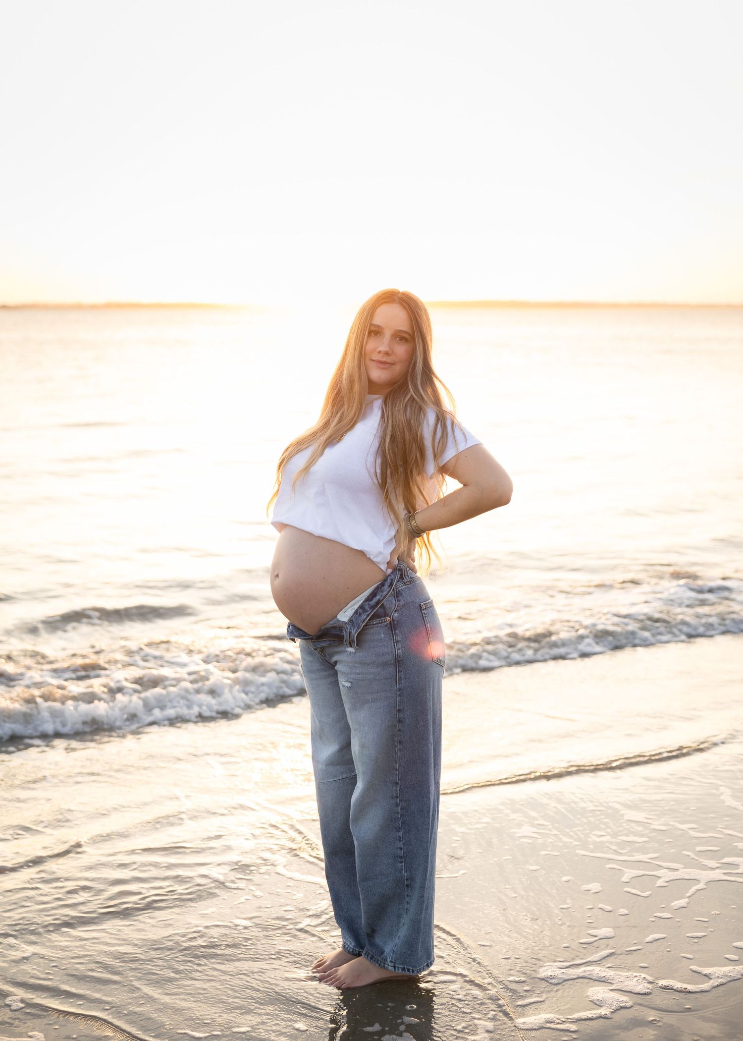 A person in jeans and white shirt poses on a sandy beach at golden hour.