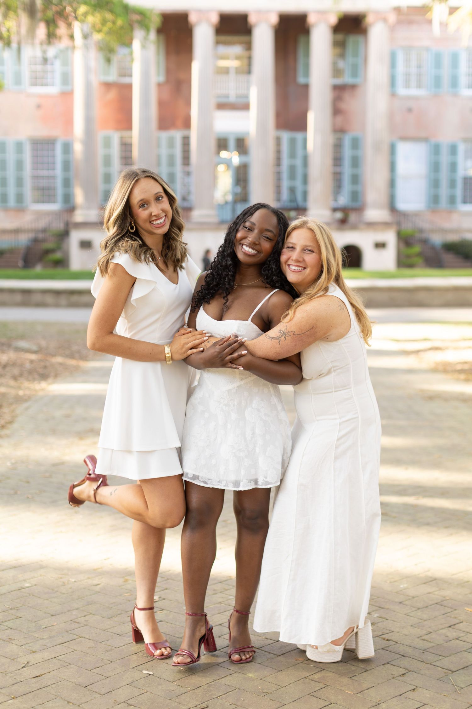 Three smiling women in white dresses embracing outdoors in front of a classic brick building.