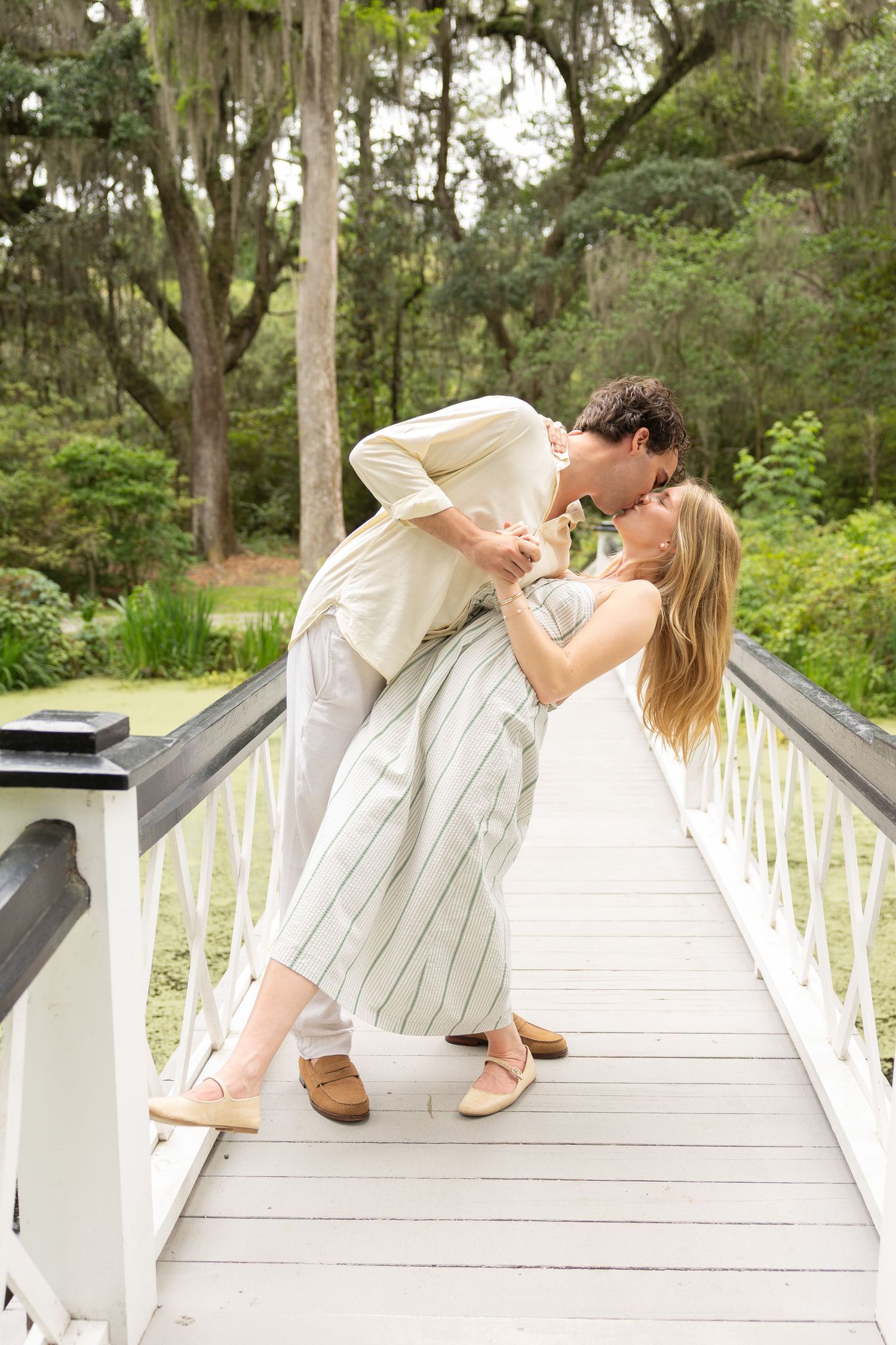 Couple sharing a romantic dip kiss on a white wooden bridge surrounded by lush green trees.