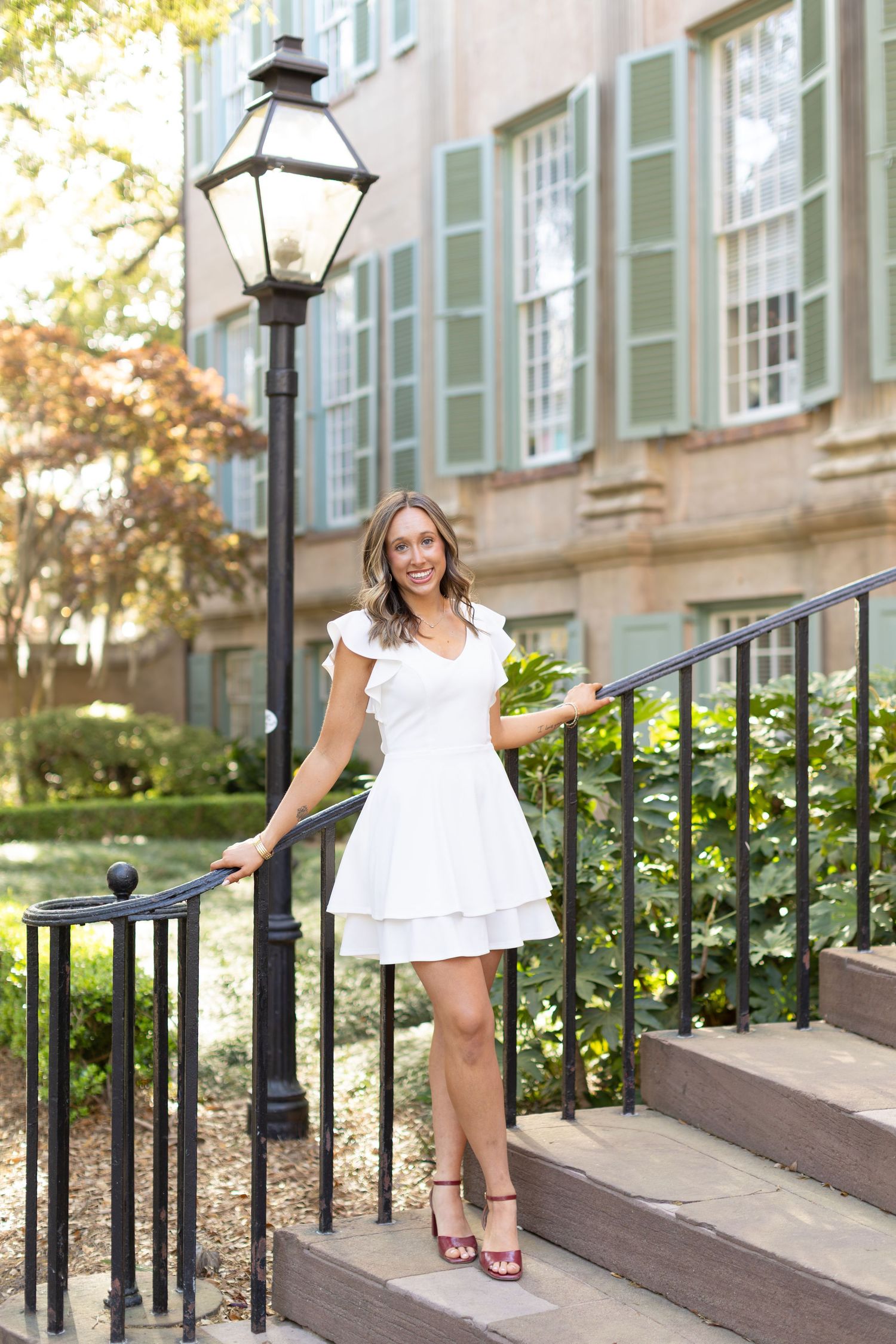 Woman in white dress posing on steps near a black lamppost outside a historic building with green shutters.