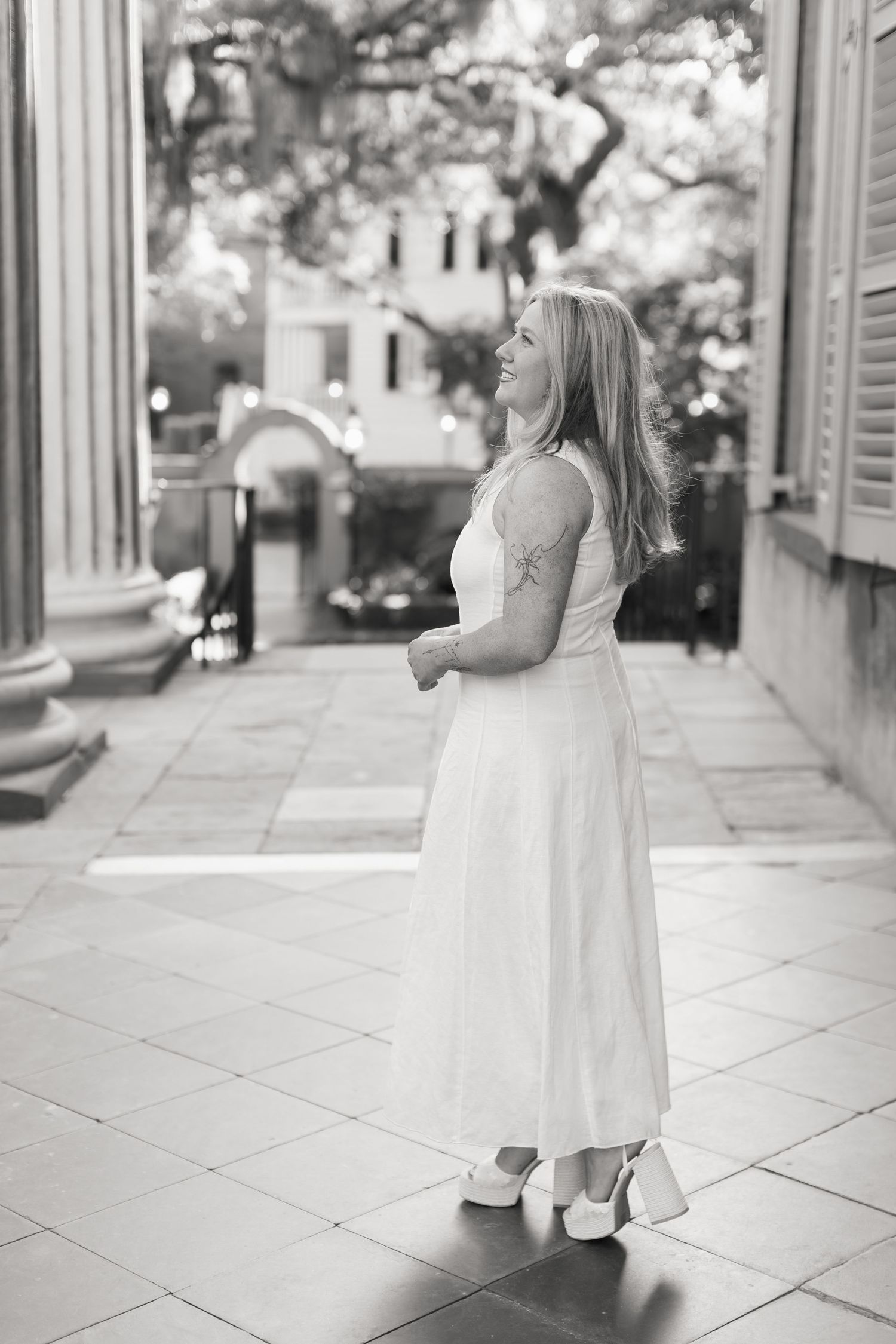 Black and white photo of a smiling blonde woman in a white maxi dress and heels standing on a tiled outdoor patio.