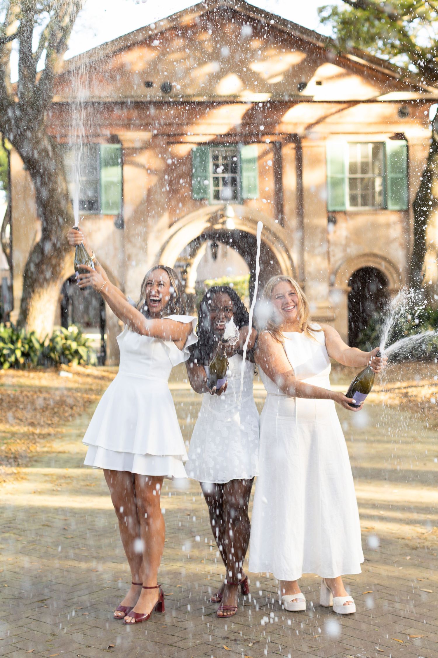 Three women in white dresses joyfully popping champagne bottles outdoors near a historic brick building.