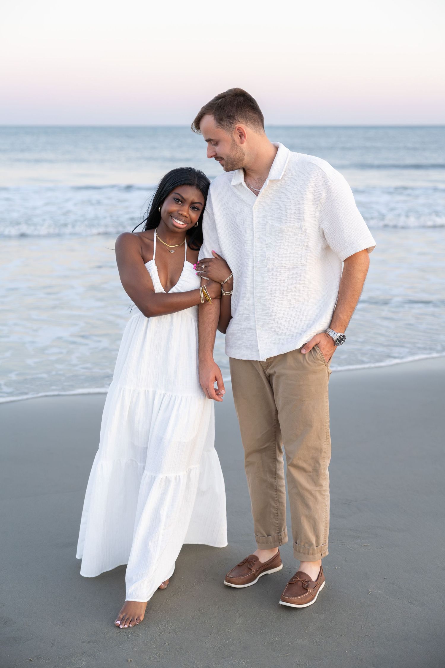Interracial couple smiling and embracing on a sandy beach at sunset, wearing white outfits with ocean waves behind them.