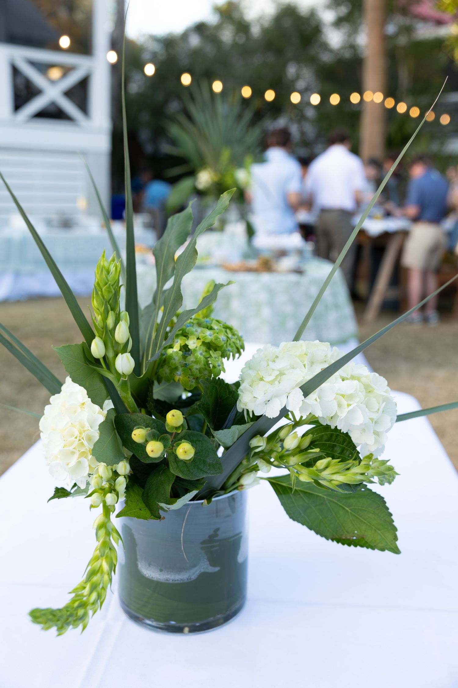 Modern white floral centerpieces with greenery in glass vases displayed on tables at an outdoor event.