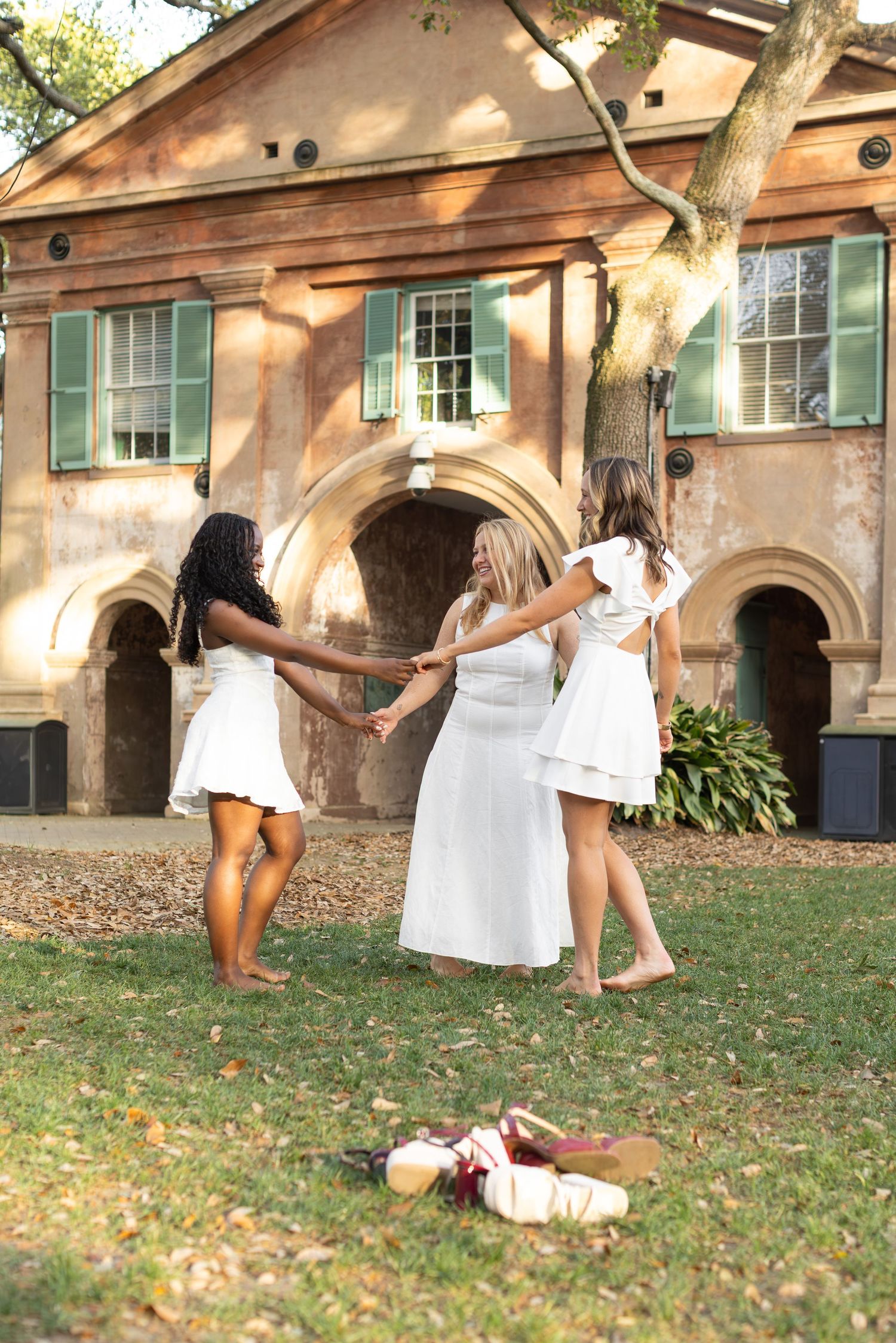 Three women in white dresses hold hands dancing on grass outside a historic brick building with green shutters.