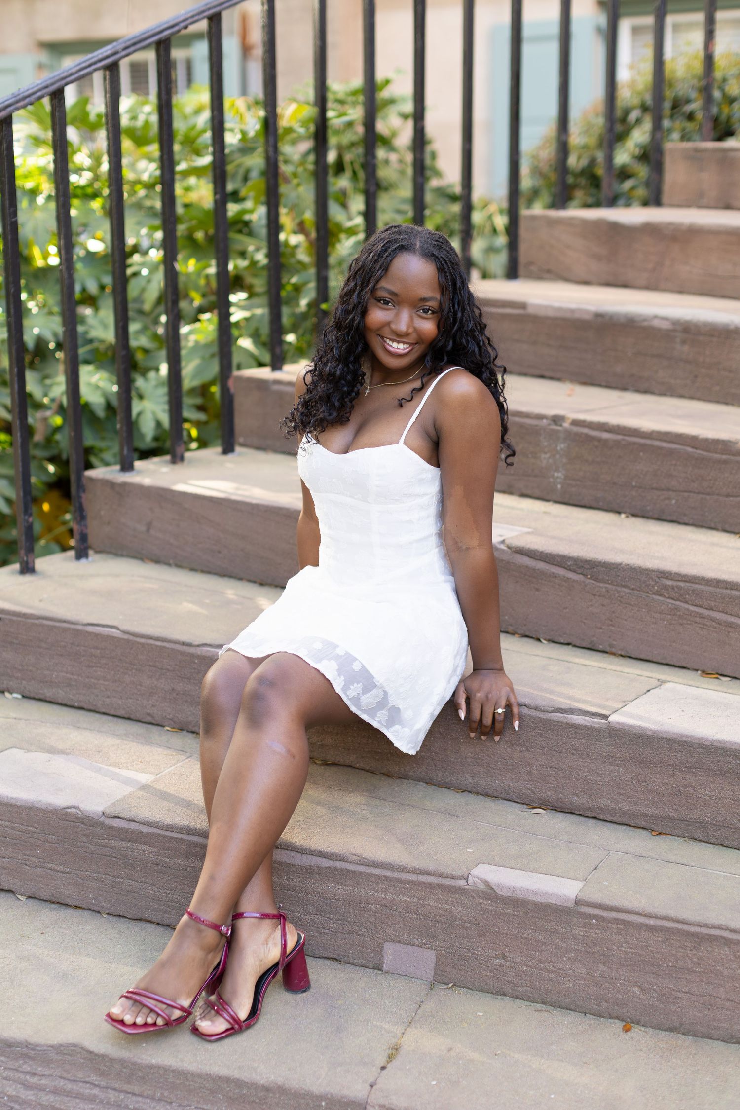 Young woman in white dress and red heels sitting on stone steps, smiling outdoors near green foliage.