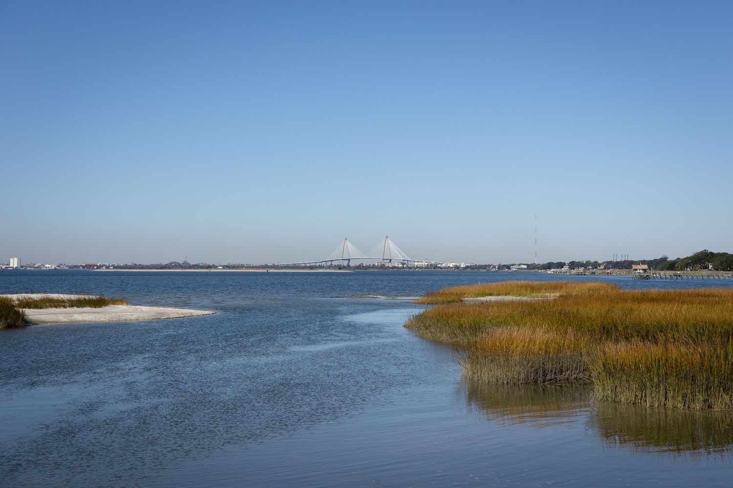 Scenic coastal marsh with golden reeds, calm blue water, and a distant bridge under a clear sky.