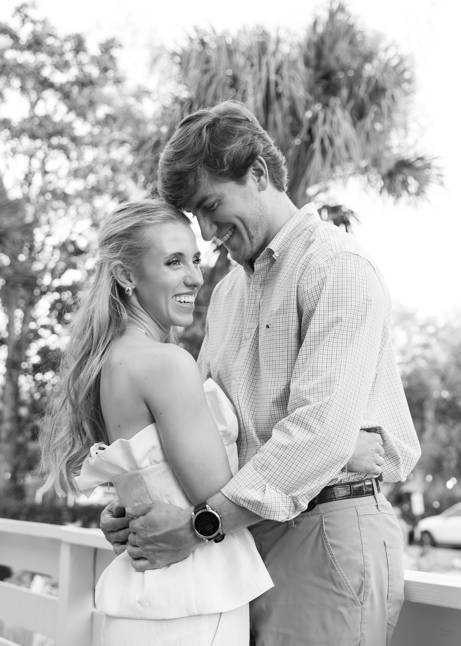 A romantic black and white photo of a couple sharing a joyful embrace on a porch with palm trees in the background.
