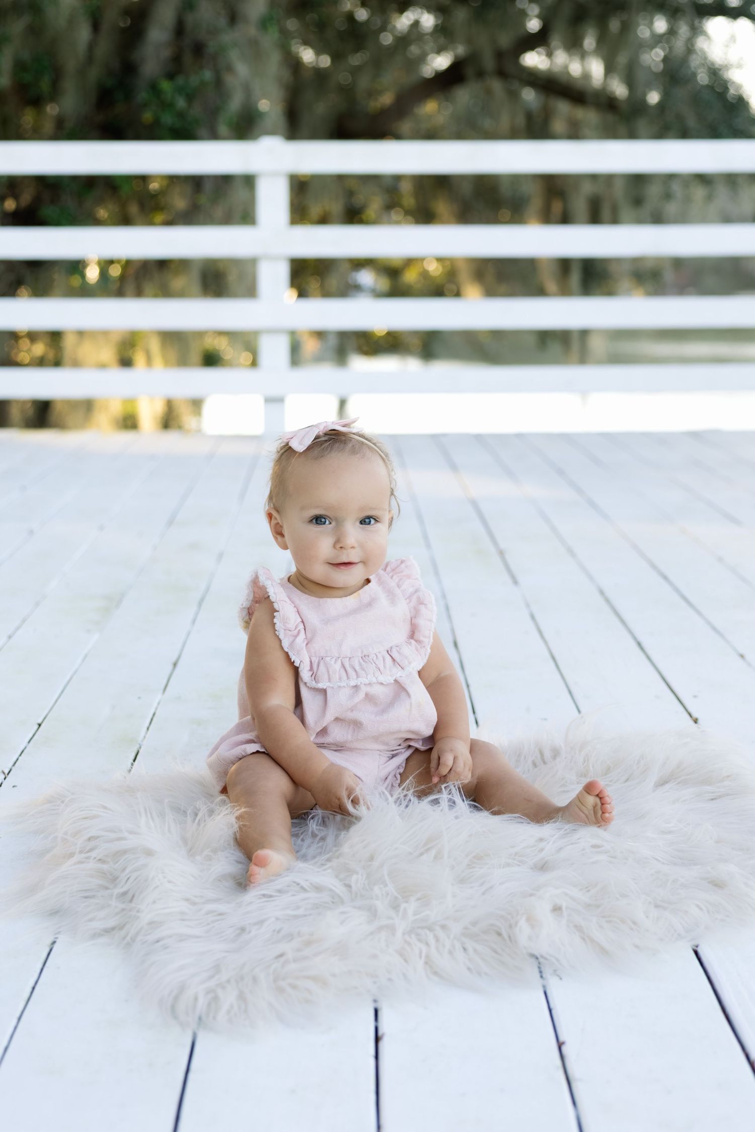 Baby in pink dress sitting on white fur rug against rustic white fence backdrop outdoors.