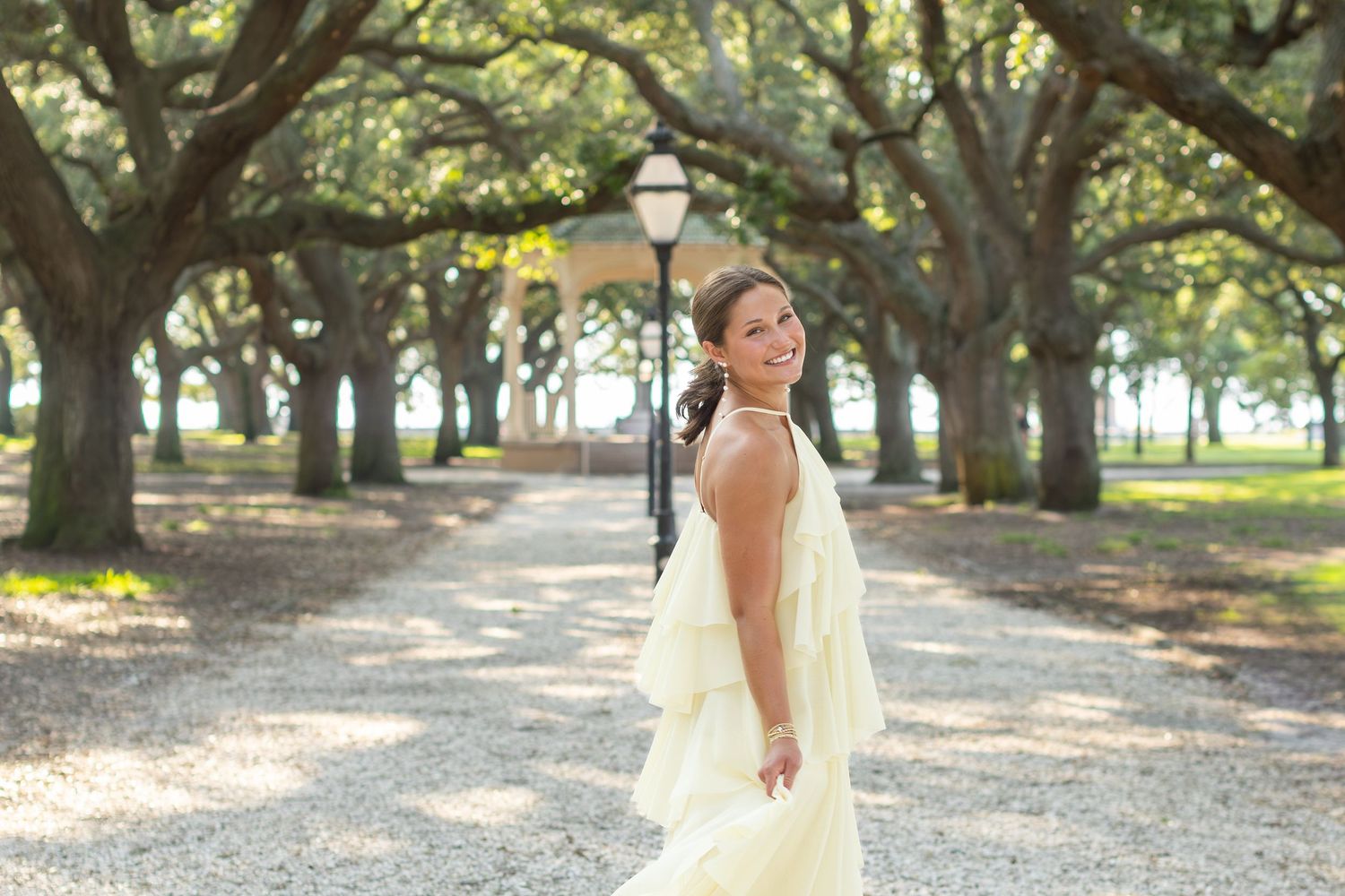 Woman in elegant backless yellow gown posing on a tree-lined gravel path with a lamp post behind her.
