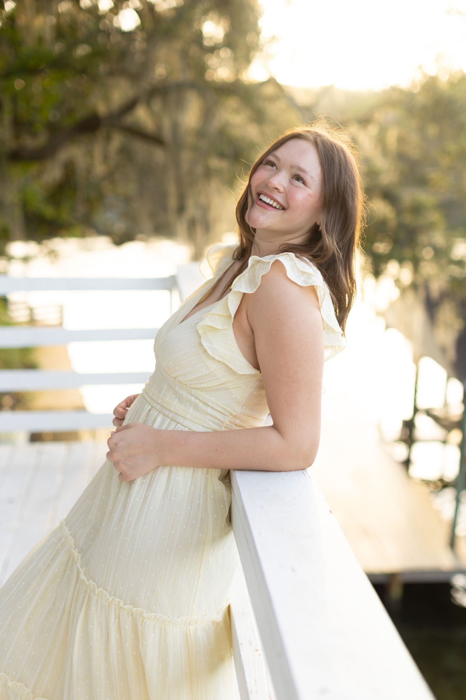 Smiling young woman in a yellow ruffle dress leaning on a white railing on a sunlit dock surrounded by trees.