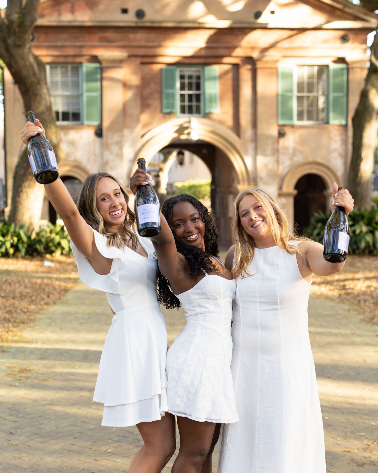 Three women in white dresses celebrate outdoors holding wine bottles in front of a brick building.