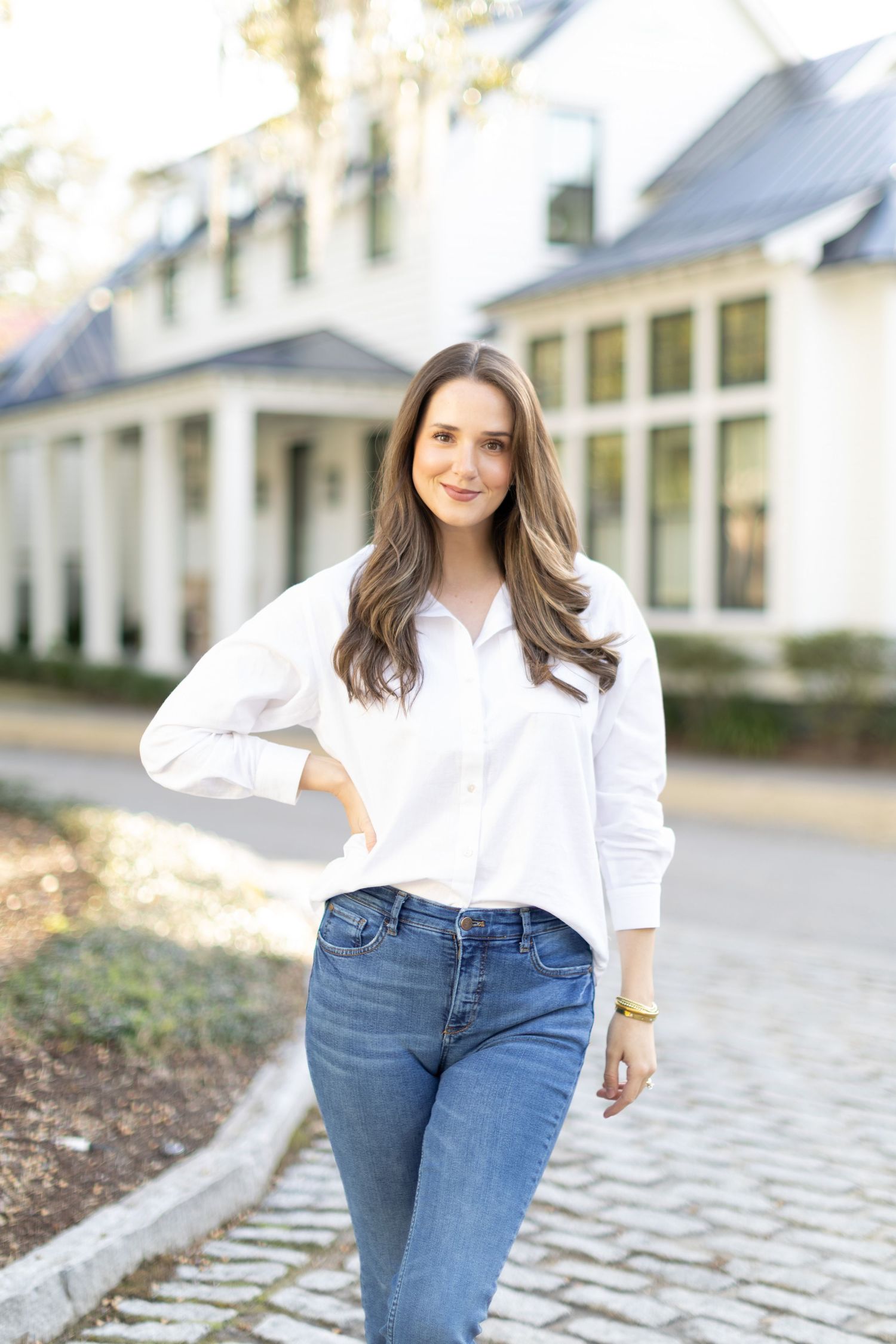 A person in a crisp white button-down shirt and blue jeans poses casually outdoors against a light-colored building.