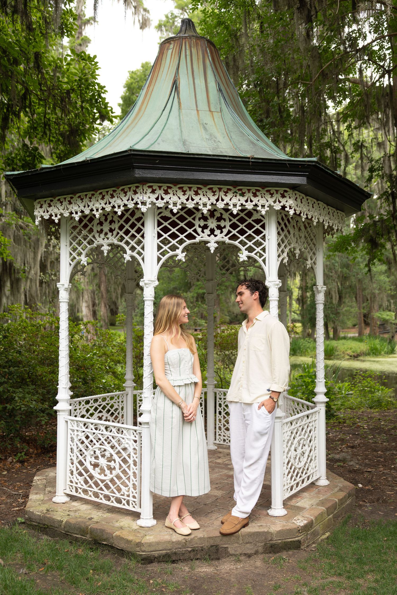Couple standing together in a white ornate gazebo surrounded by lush green trees draped with Spanish moss.