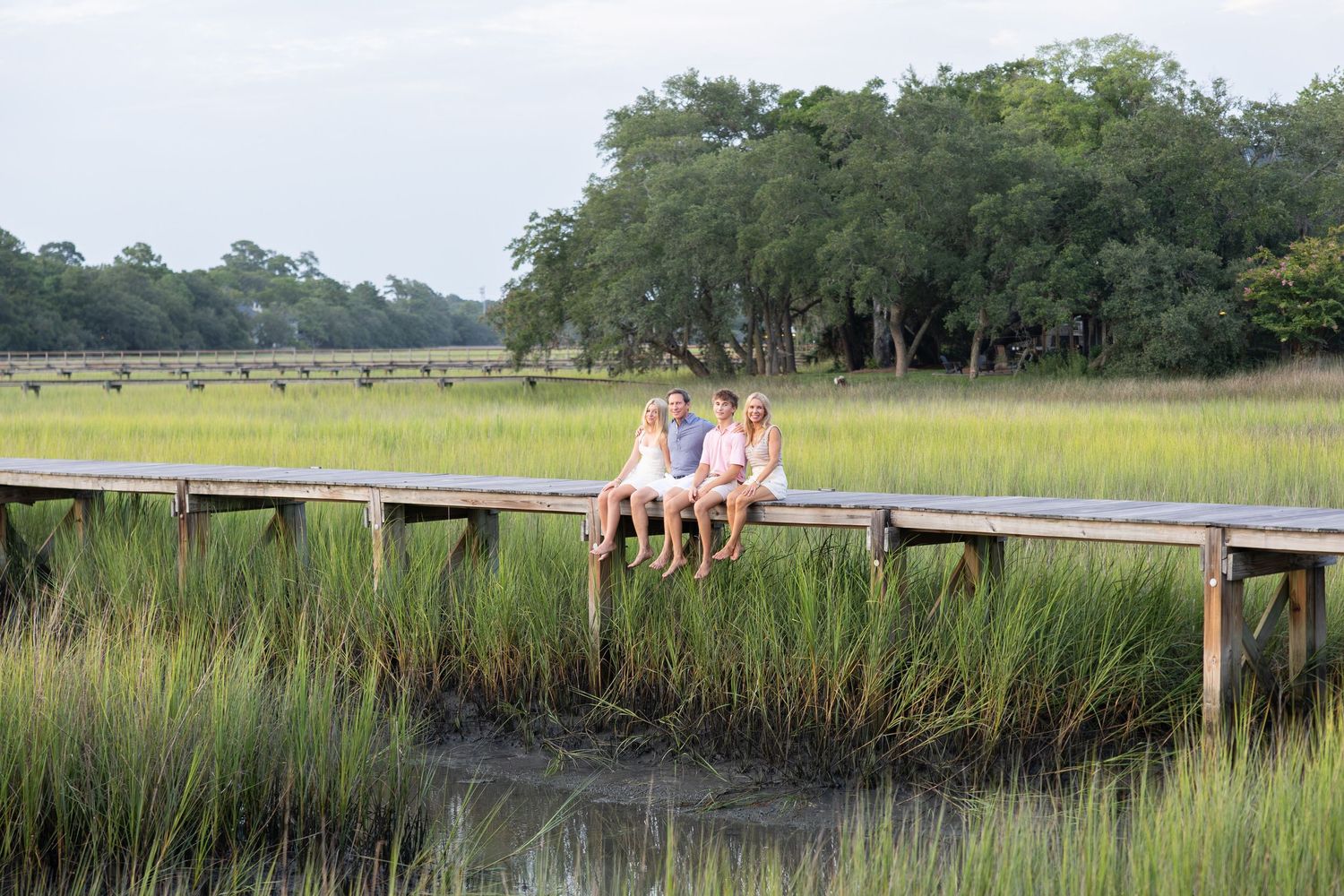 Three children sit on a wooden dock overlooking a lush green salt marsh with trees in the background.