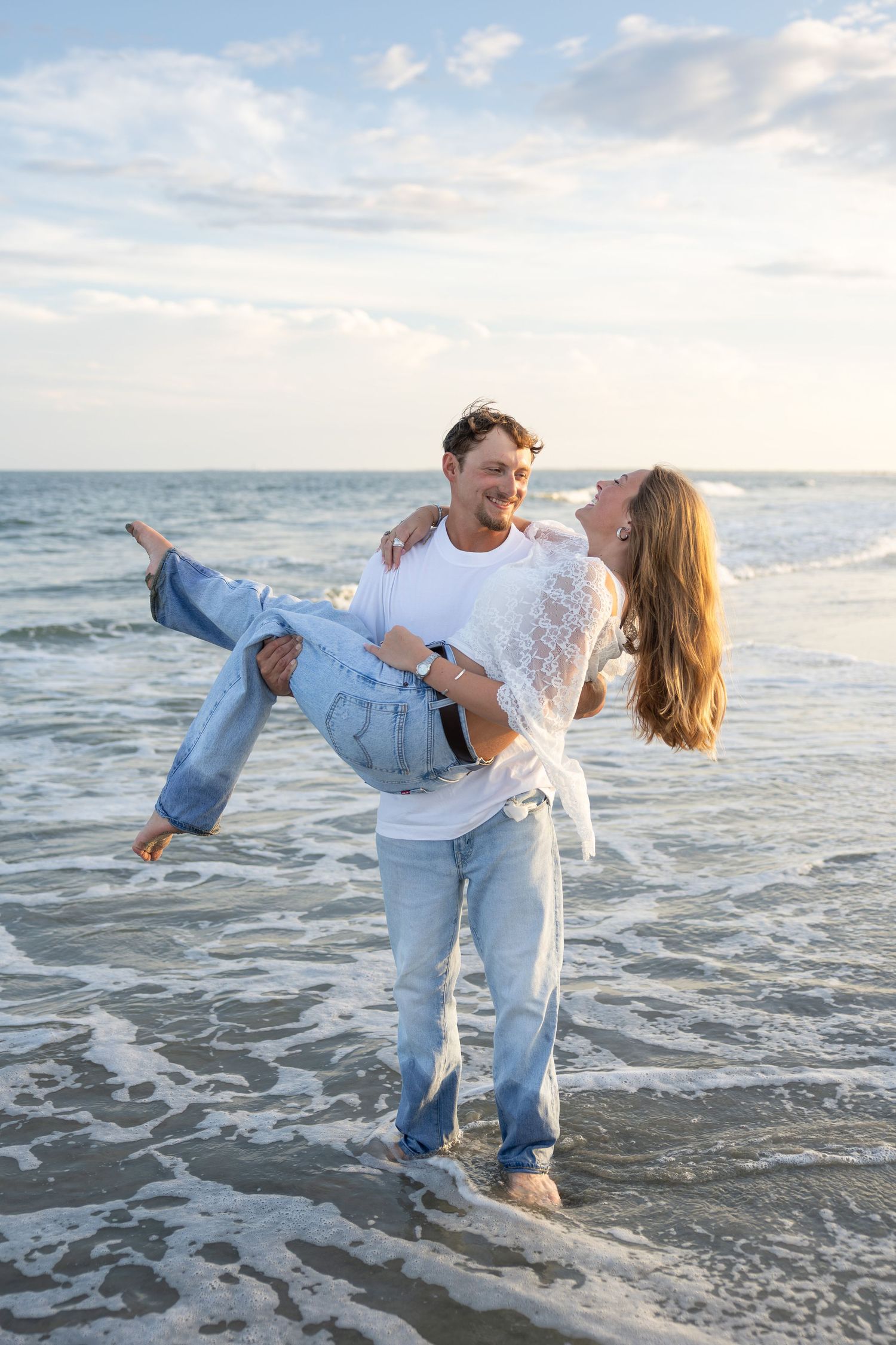 Happy couple on beach, man carrying woman in his arms, both wearing jeans, smiling at each other by ocean waves.