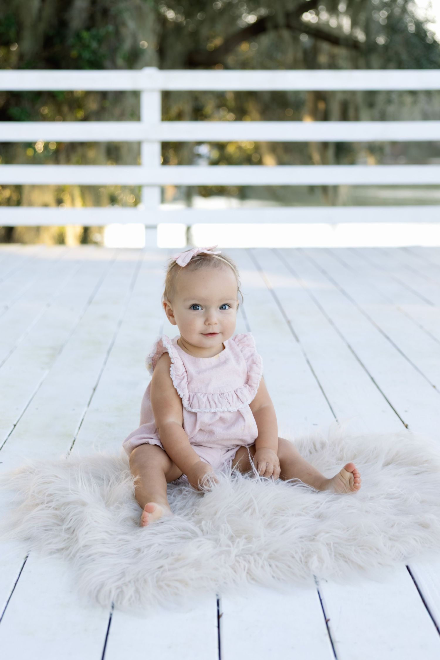 Small child wearing pink ruffled romper sits on fluffy white rug on wooden deck with white railing in background.