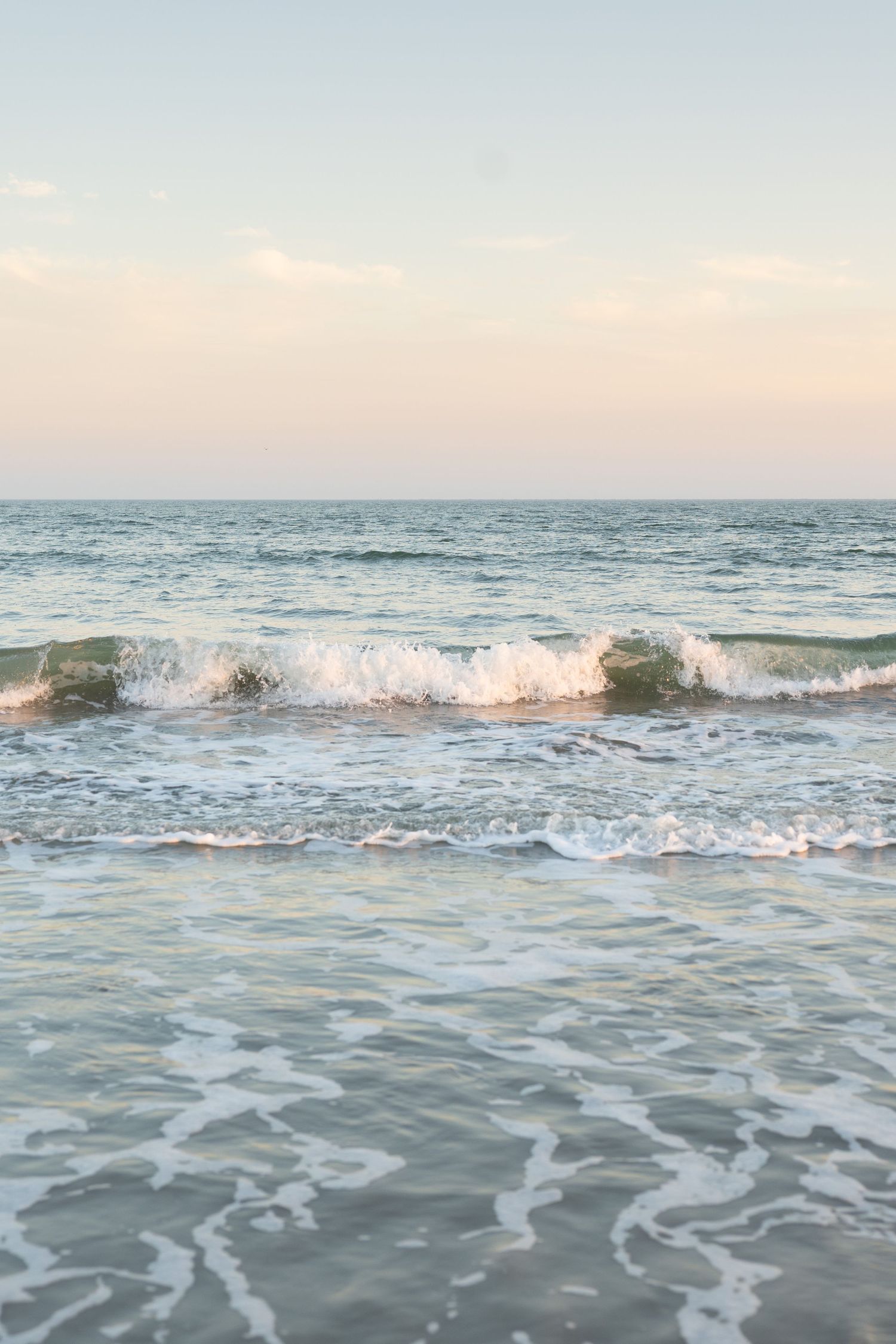 Ocean waves roll onto a beach during sunset with soft pastel colors reflecting in the foamy water.