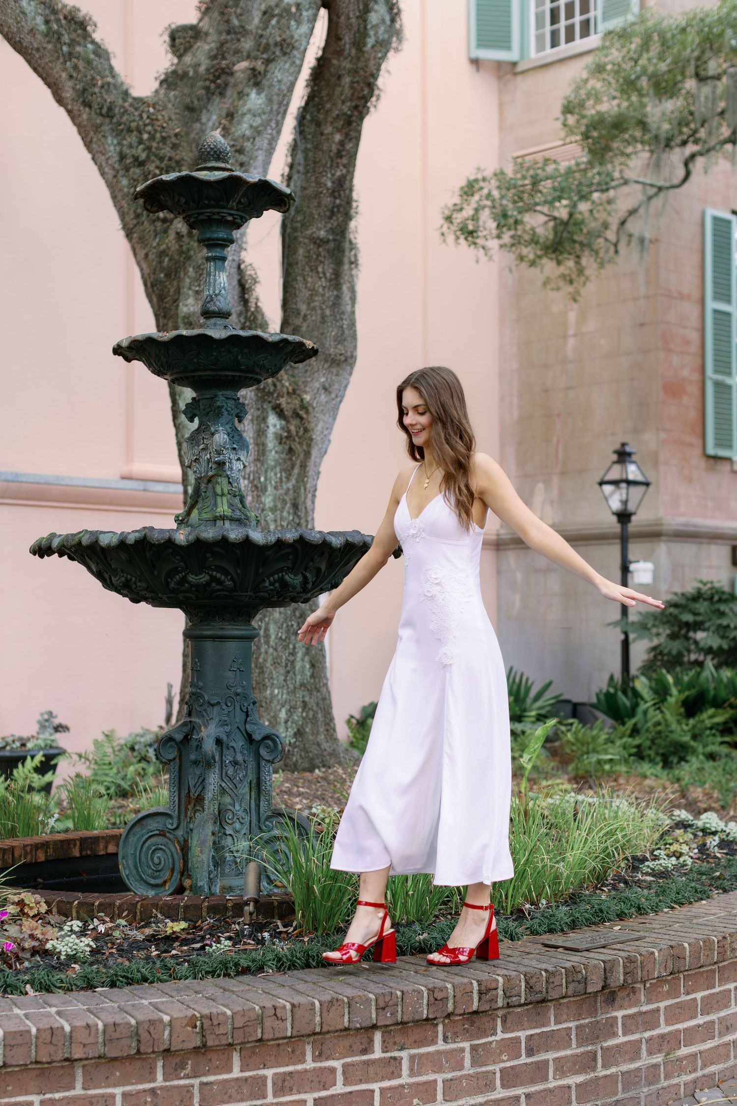A figure in a white dress and red shoes balances gracefully along a brick wall near ornate lamp posts.