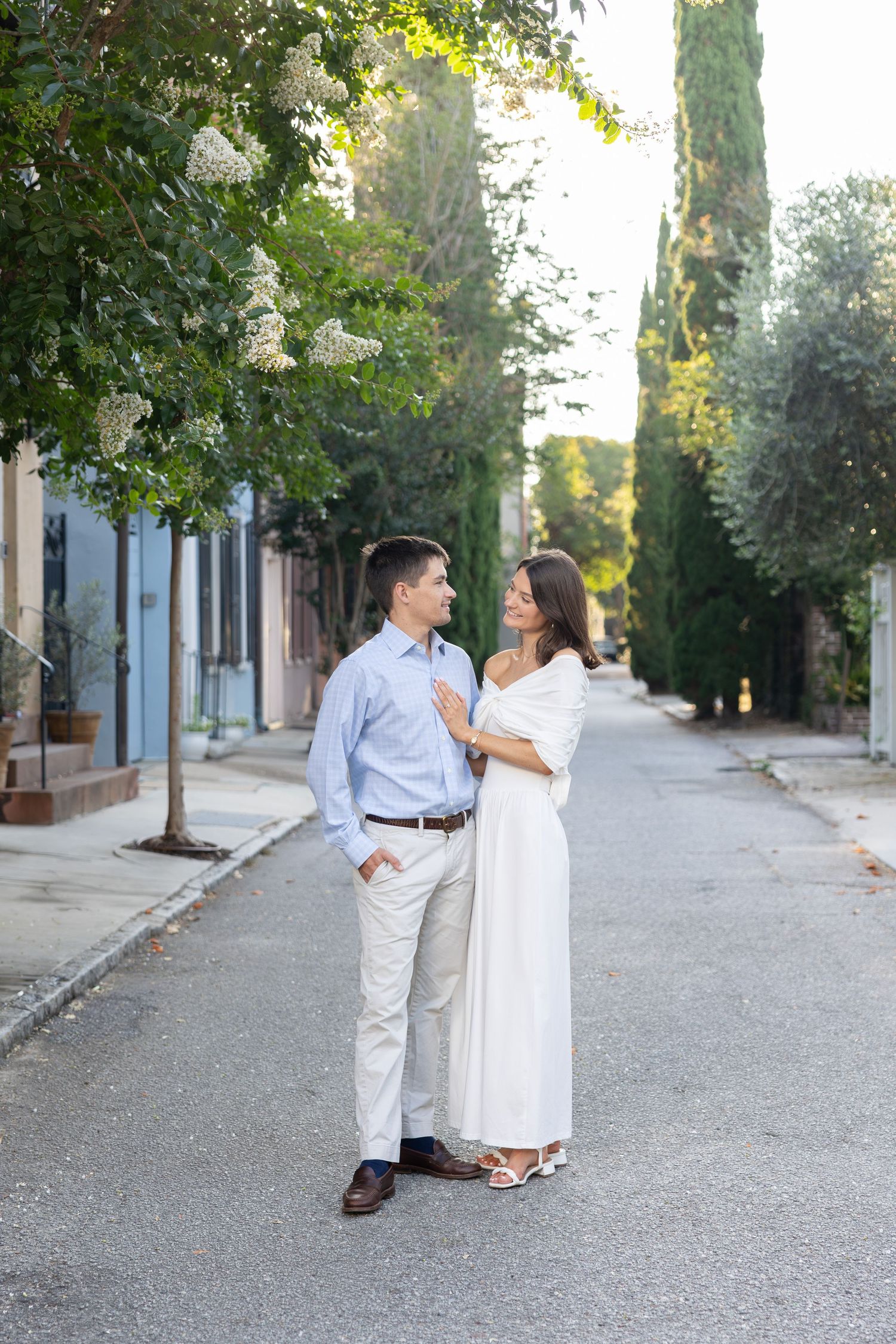 Couple in elegant attire embracing on a quiet street lined with cypress trees and blooming flowers in warm sunlight.