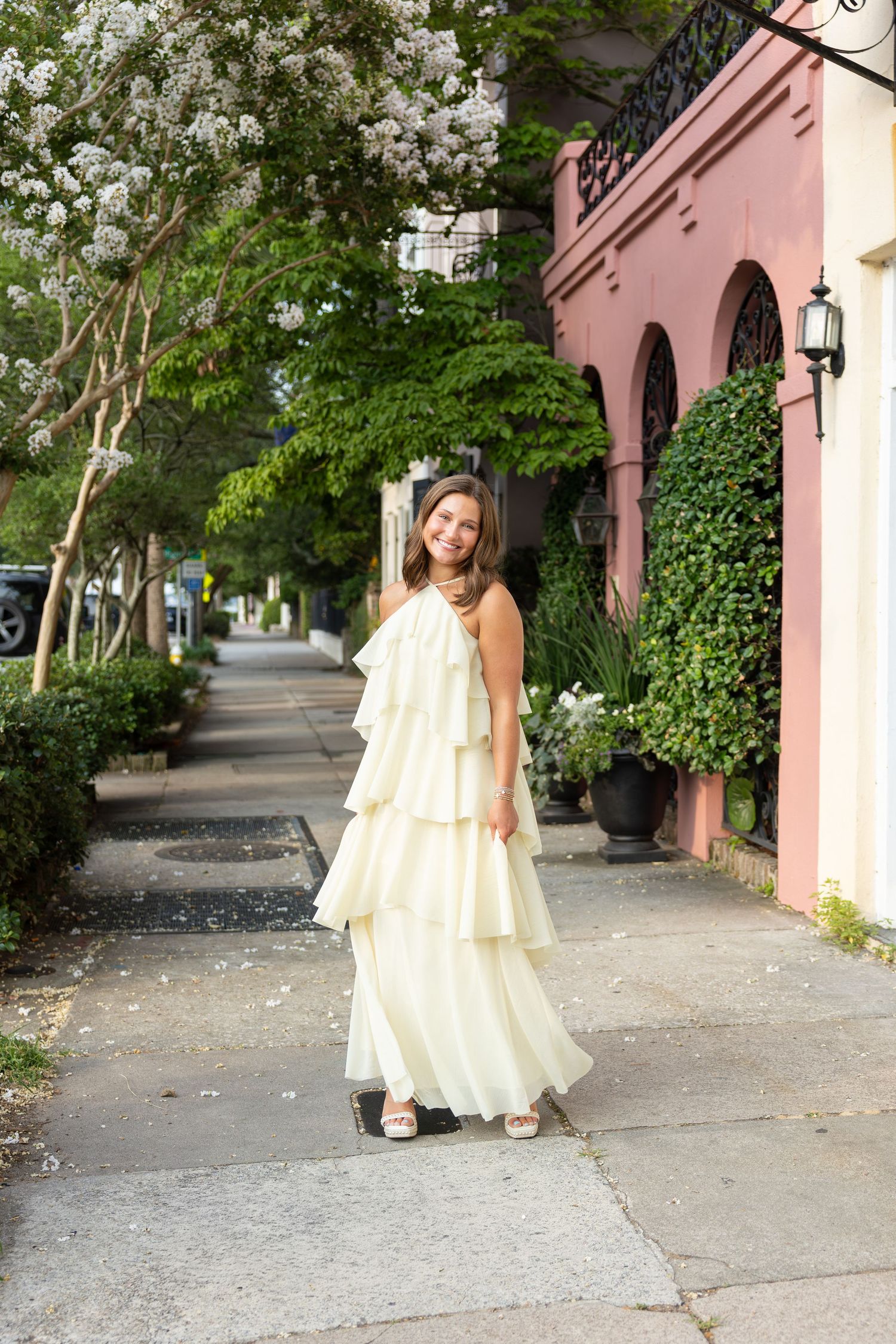 Young girl in white tiered ruffle gown posing on a charming sidewalk with pink building and lush greenery.