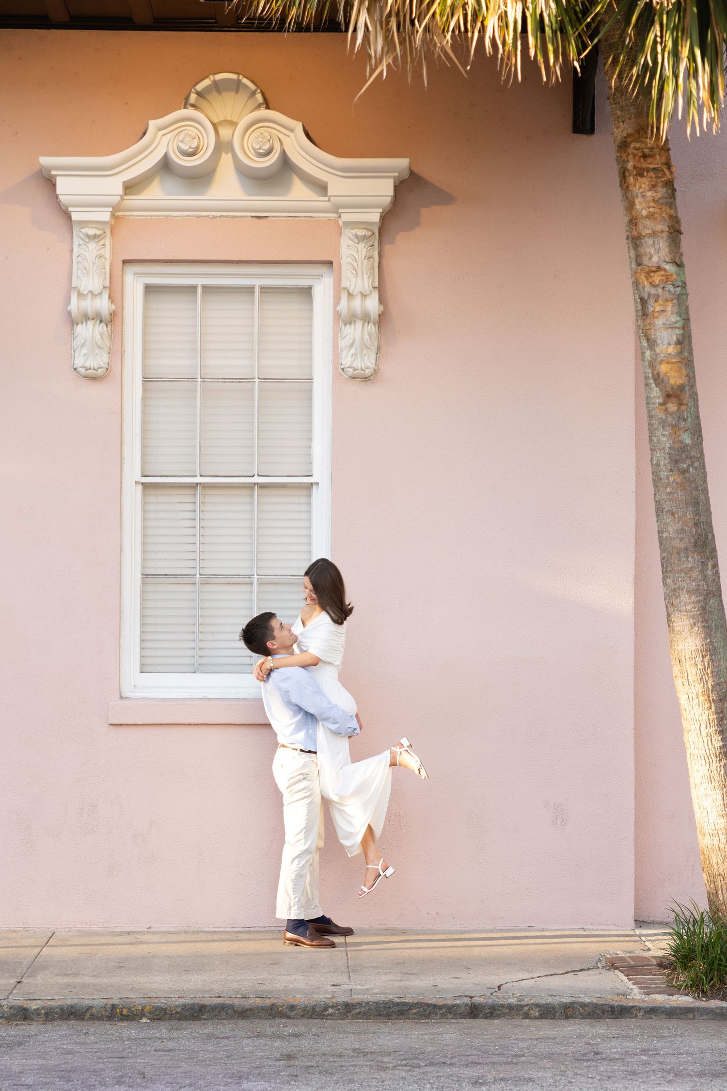 A man lifts a woman joyfully in front of a pink building with ornate window trim and a palm tree.