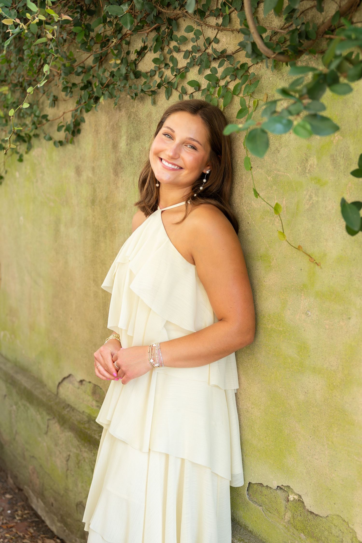 Smiling young woman in white ruffled halter dress posing against mossy wall with green ivy backdrop.