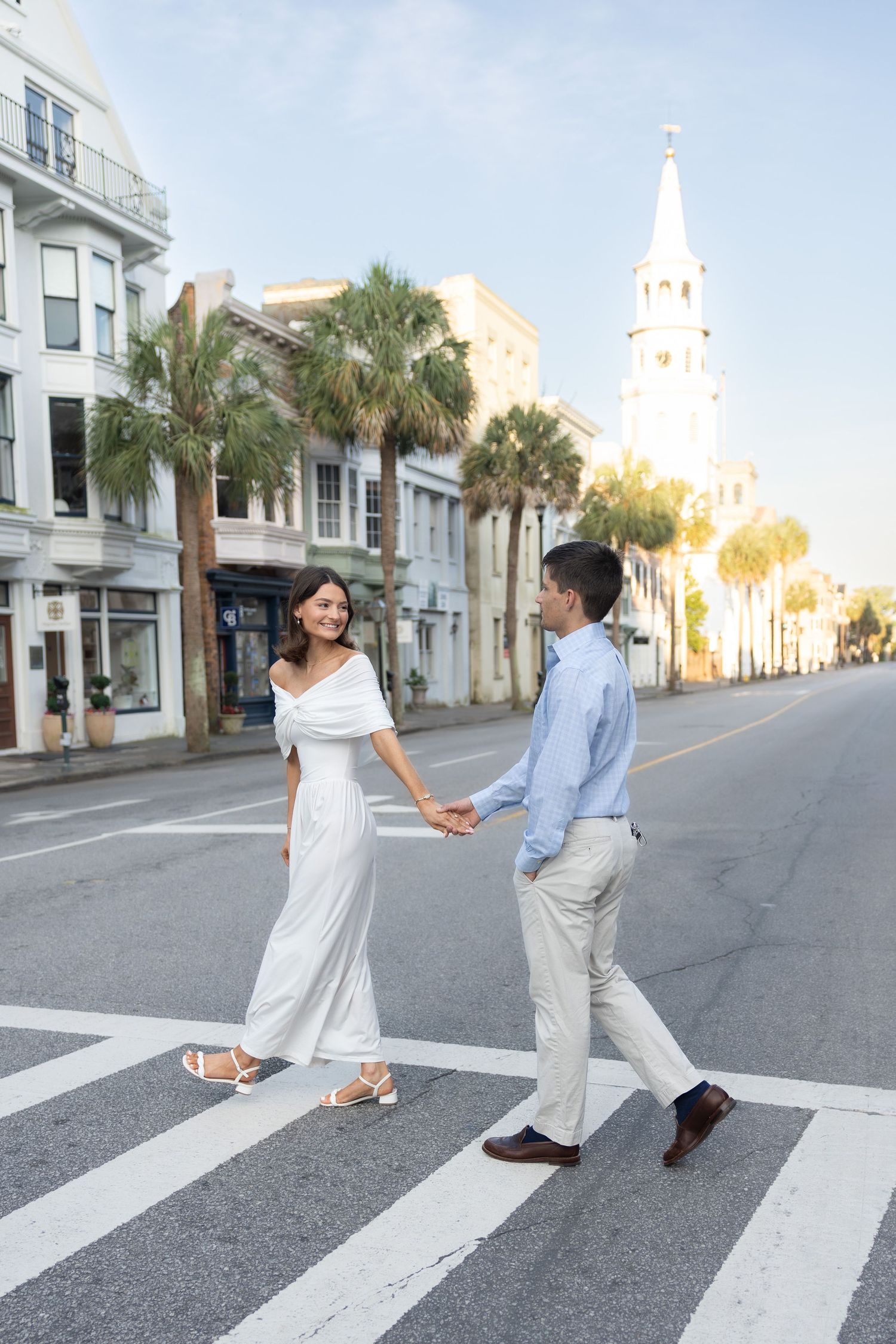 Couple holding hands crossing a sunny street in Charleston, SC, with palm trees and a white church steeple in the background.