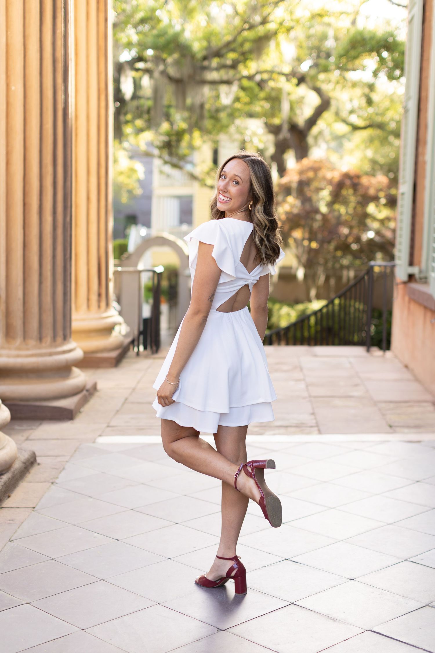 Young woman in white ruffled dress and red heels posing outdoors near classical columns in a sunny courtyard.