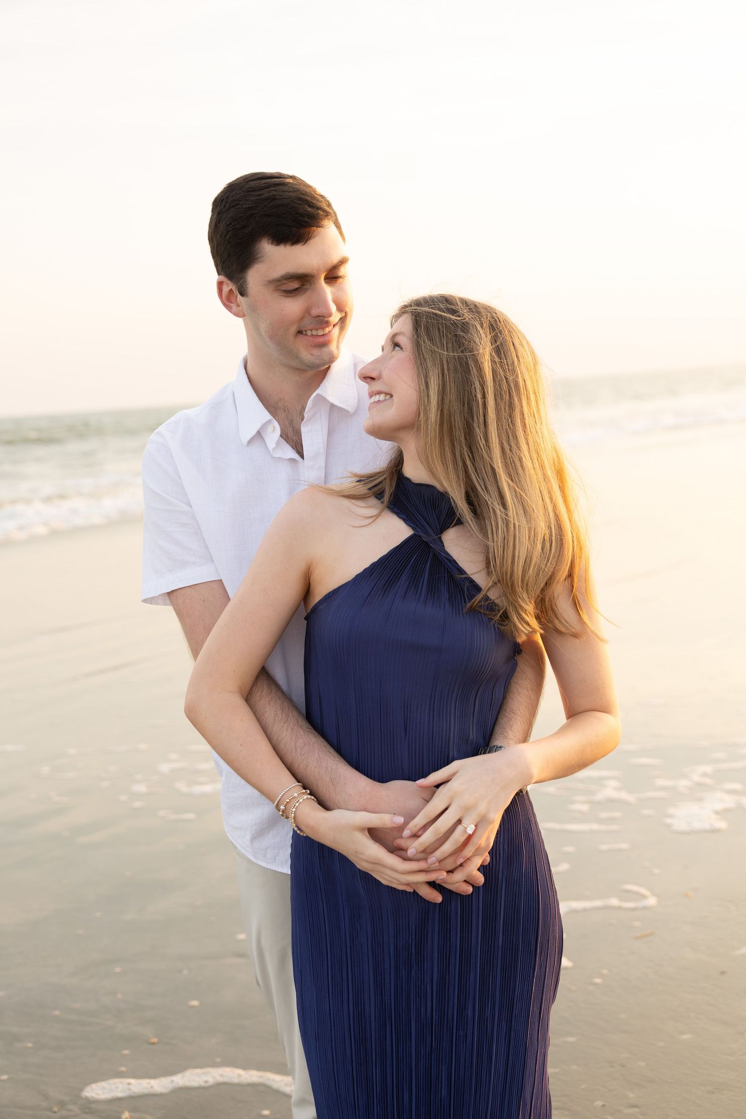 A couple embraces on a serene beach at sunset, with the woman wearing an elegant navy halter dress.