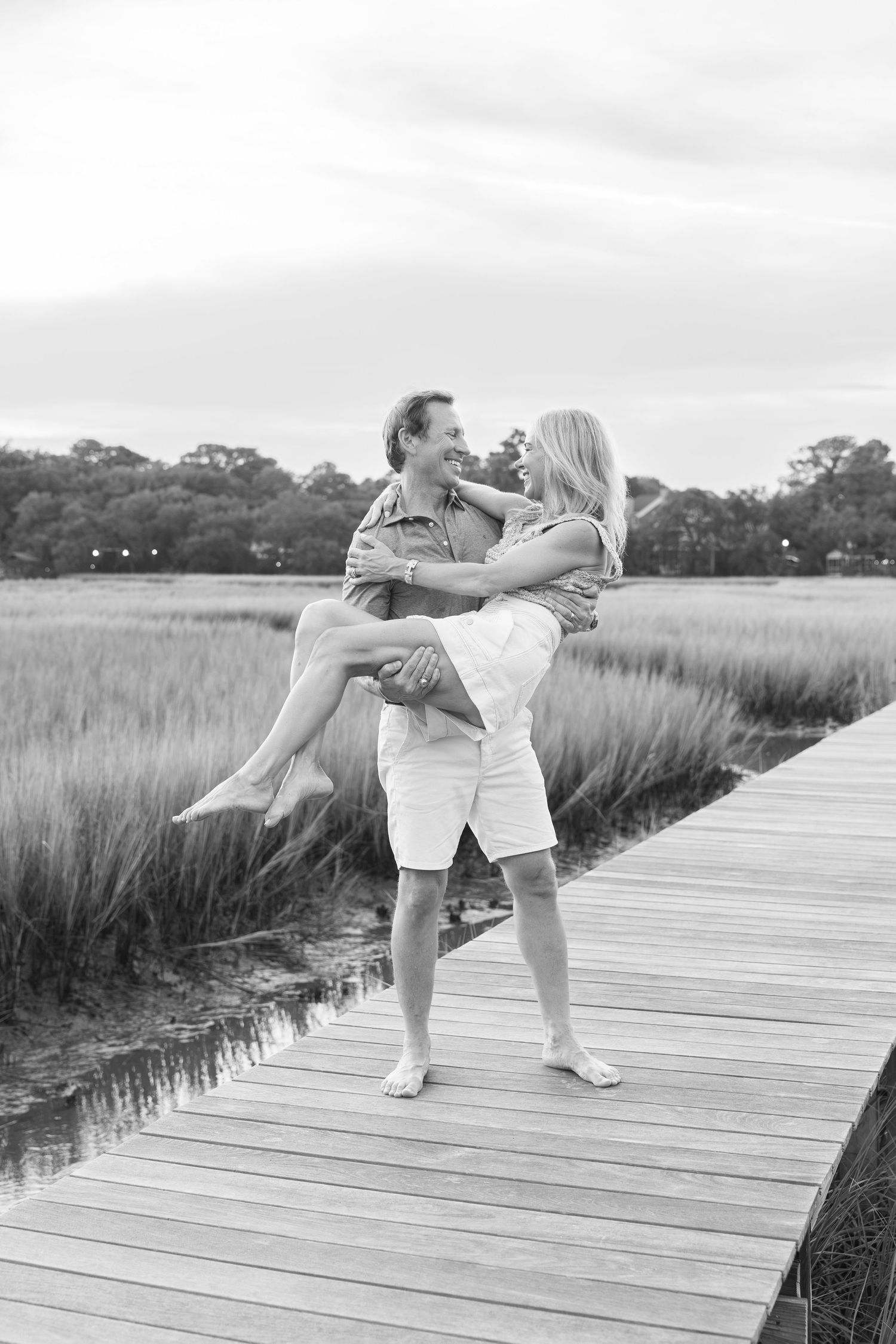 Black and white photo of a man lifting a woman on a wooden dock surrounded by marsh grass.