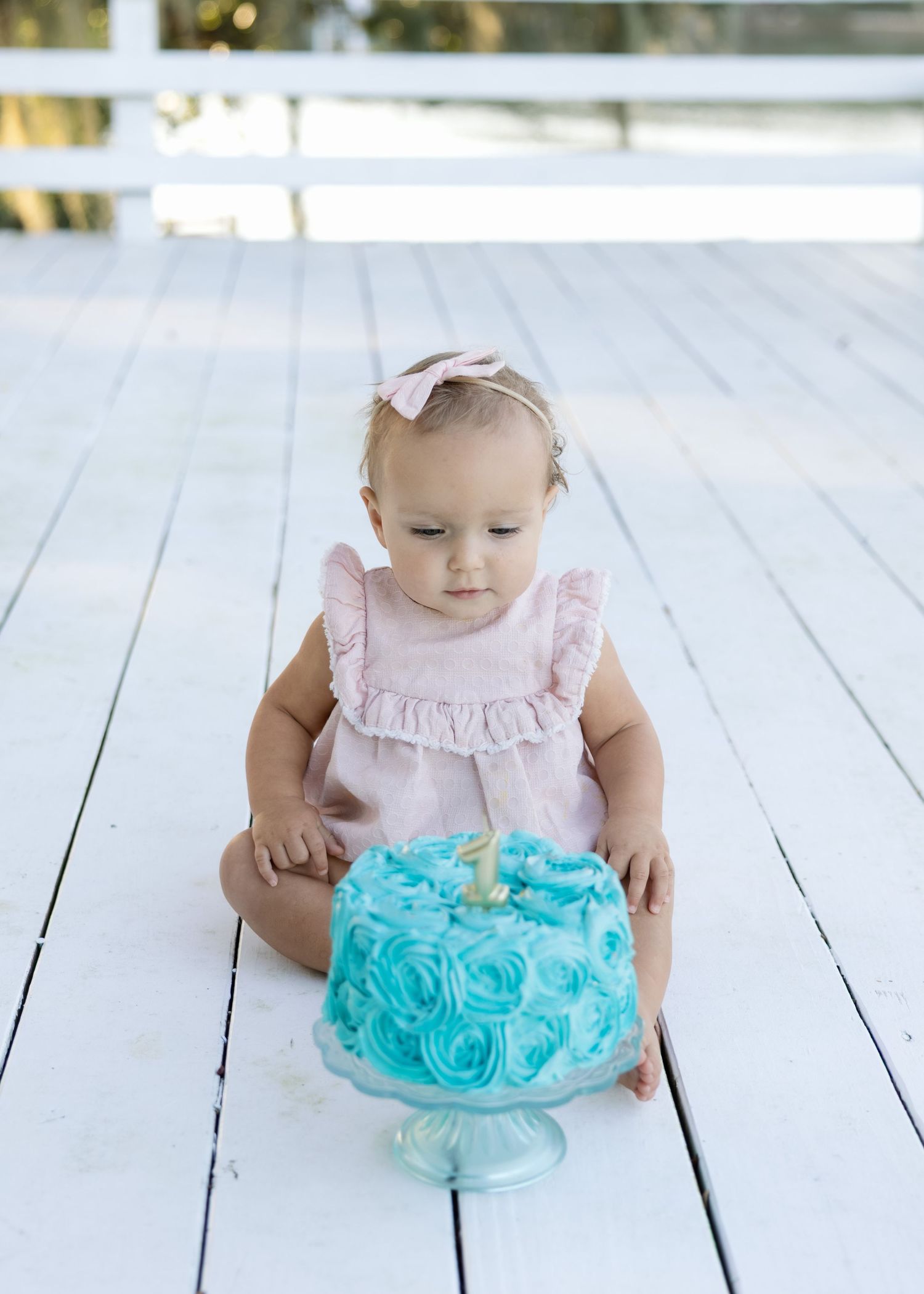 Baby girl in pink dress with bow headband examines a teal rosette smash cake during her first birthday photo session.