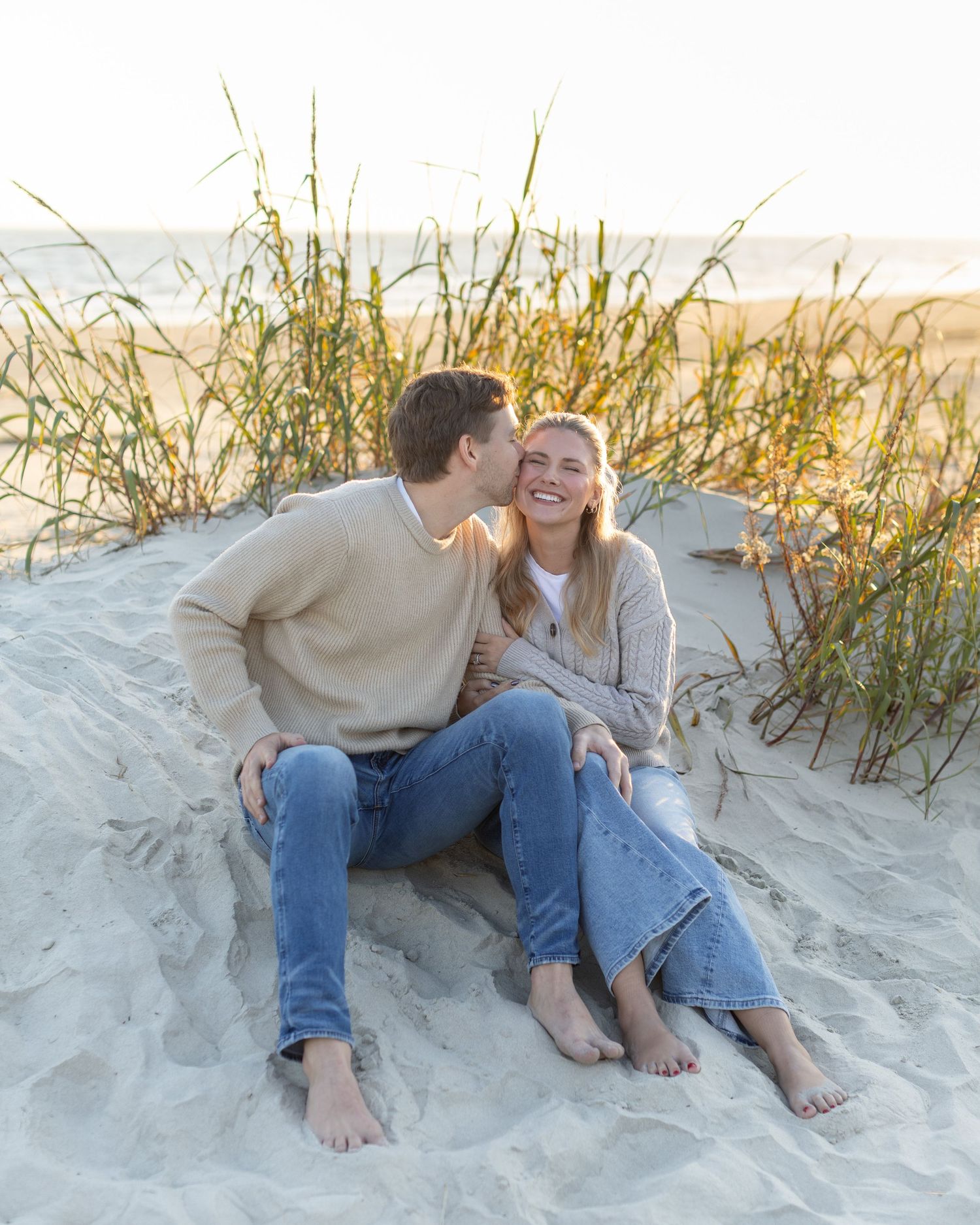 A couple shares a tender moment while sitting among beach grass on sandy dunes.