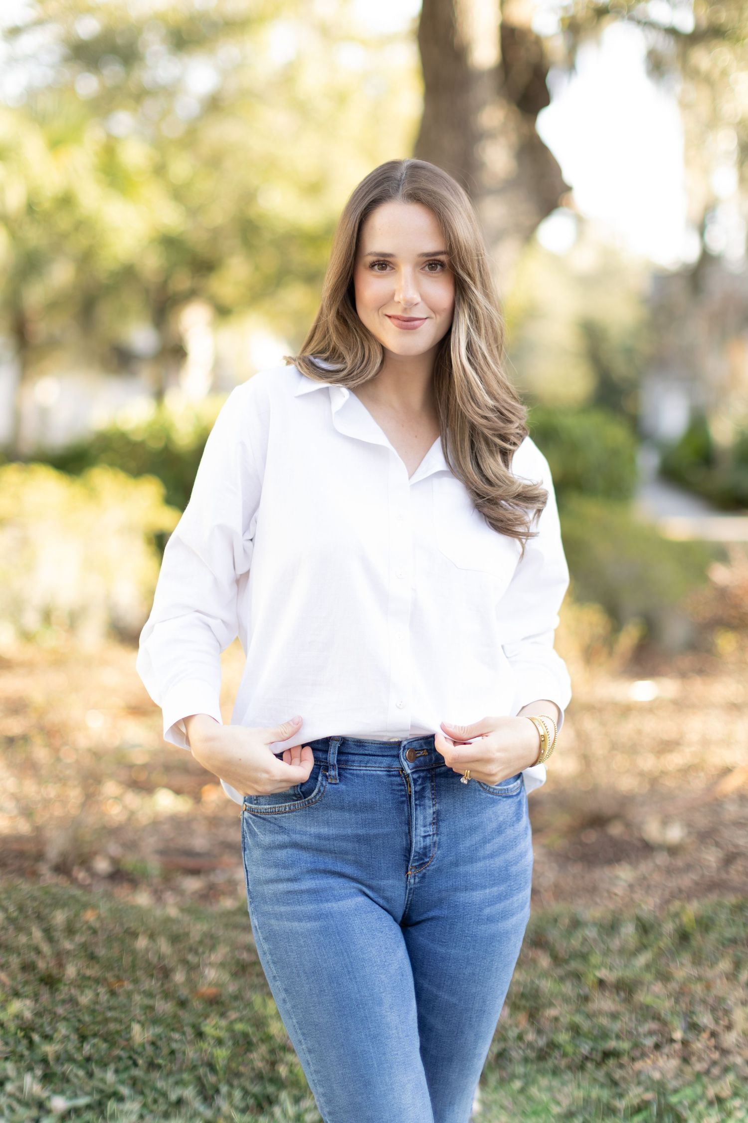 A person in a classic white button-down shirt and blue jeans poses outdoors on an autumn day with blurred foliage background.