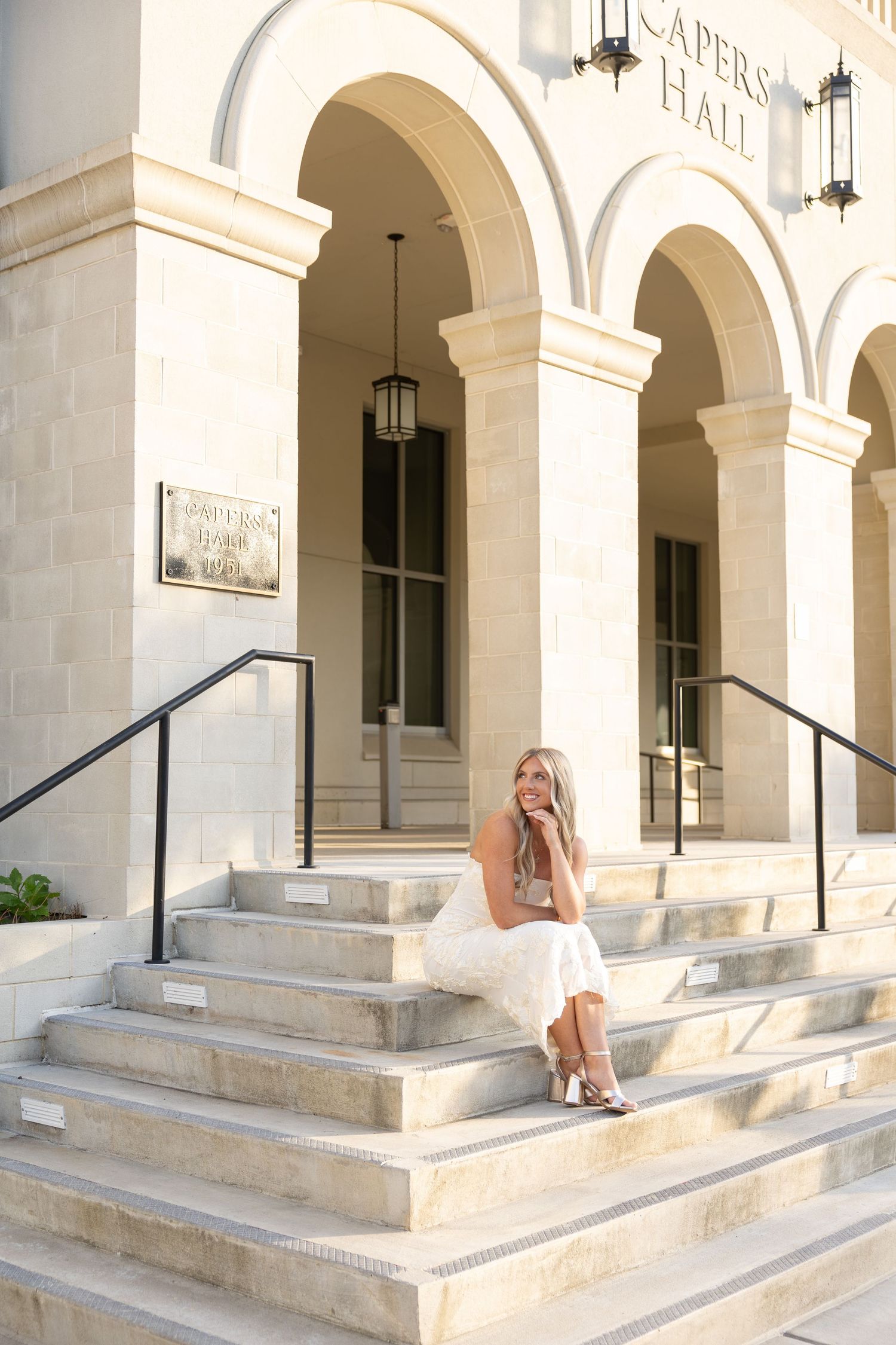 Blonde woman in white dress sitting on steps of Capers Hall building with arched columns.
