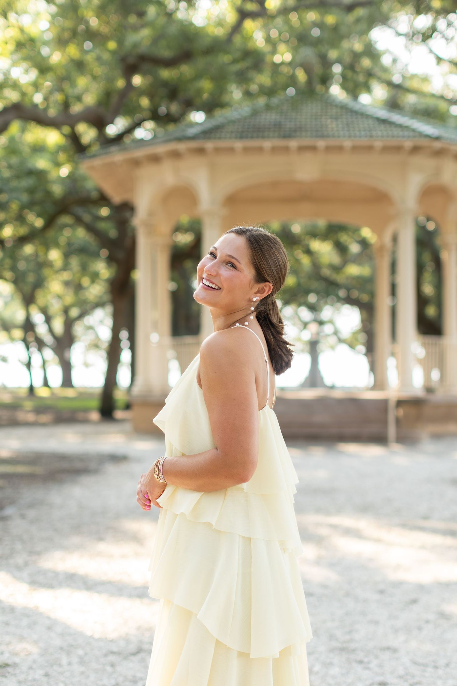Smiling woman in yellow ruffled dress poses near a gazebo surrounded by trees in a park setting.