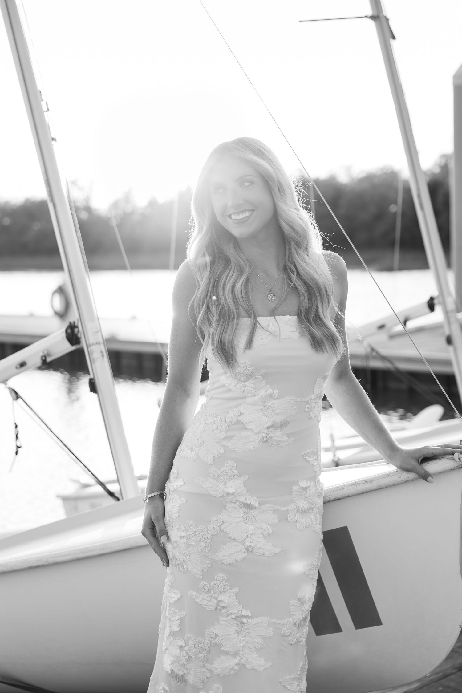 Black and white photo of a smiling woman in a floral dress standing by a sailboat at a sunny marina.