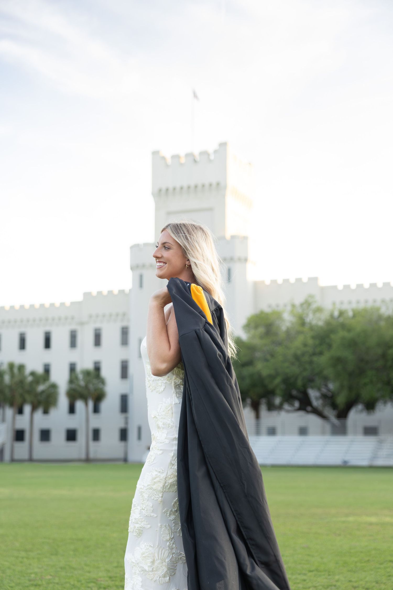 Smiling blonde graduate holding black and gold gown in white dress outside castle building on green lawn.