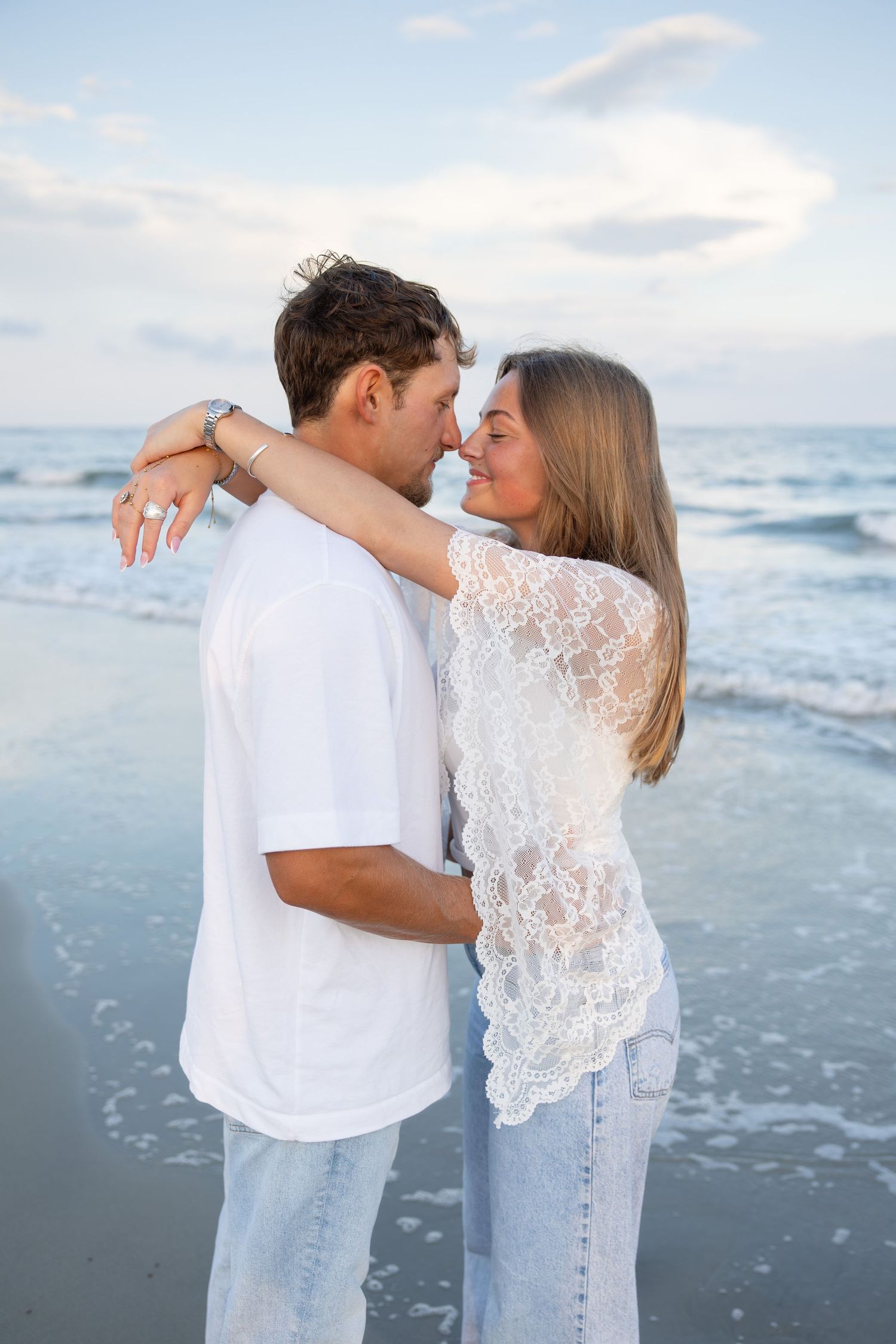 A romantic couple embraces on a beach at sunset, wearing white tops and jeans, sharing an intimate moment by the ocean waves.