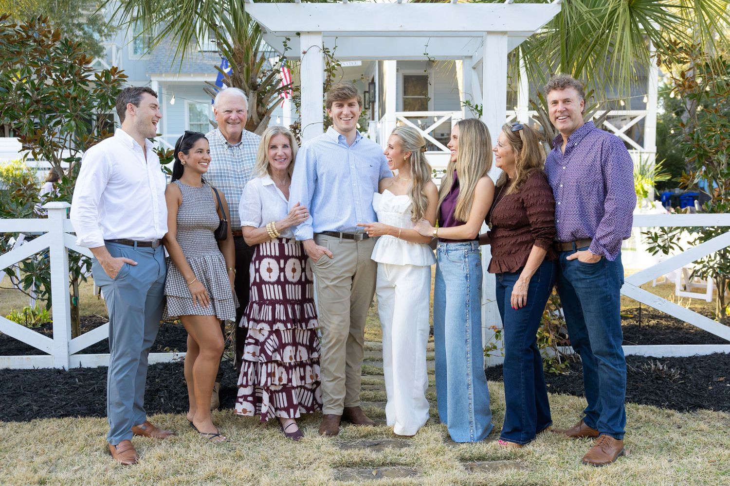 Group of people gathered outdoors near white fencing and palm trees at a casual event.