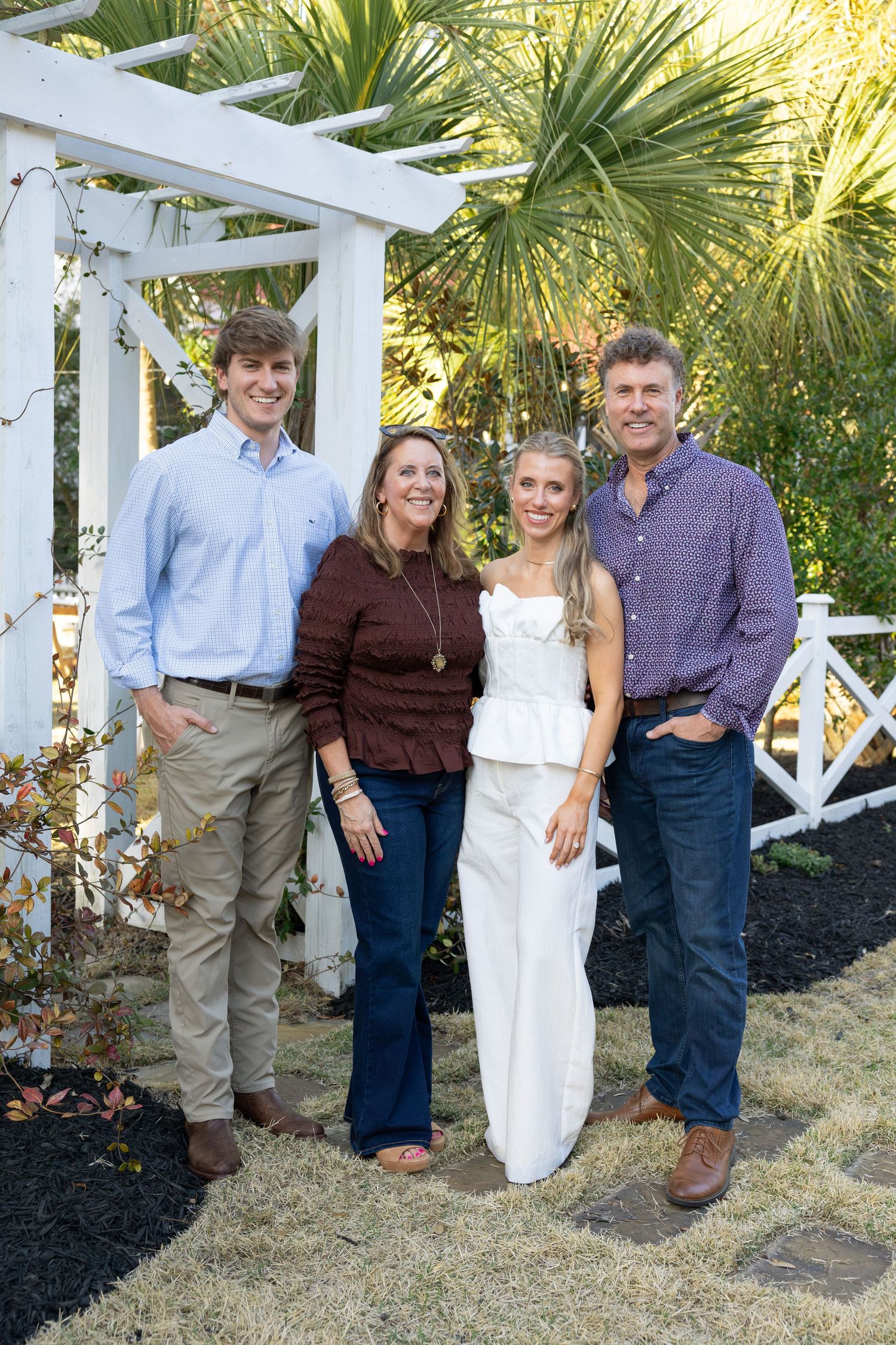 Group photo of wedding guests posing together against white fence and tropical landscaping at sunset.
