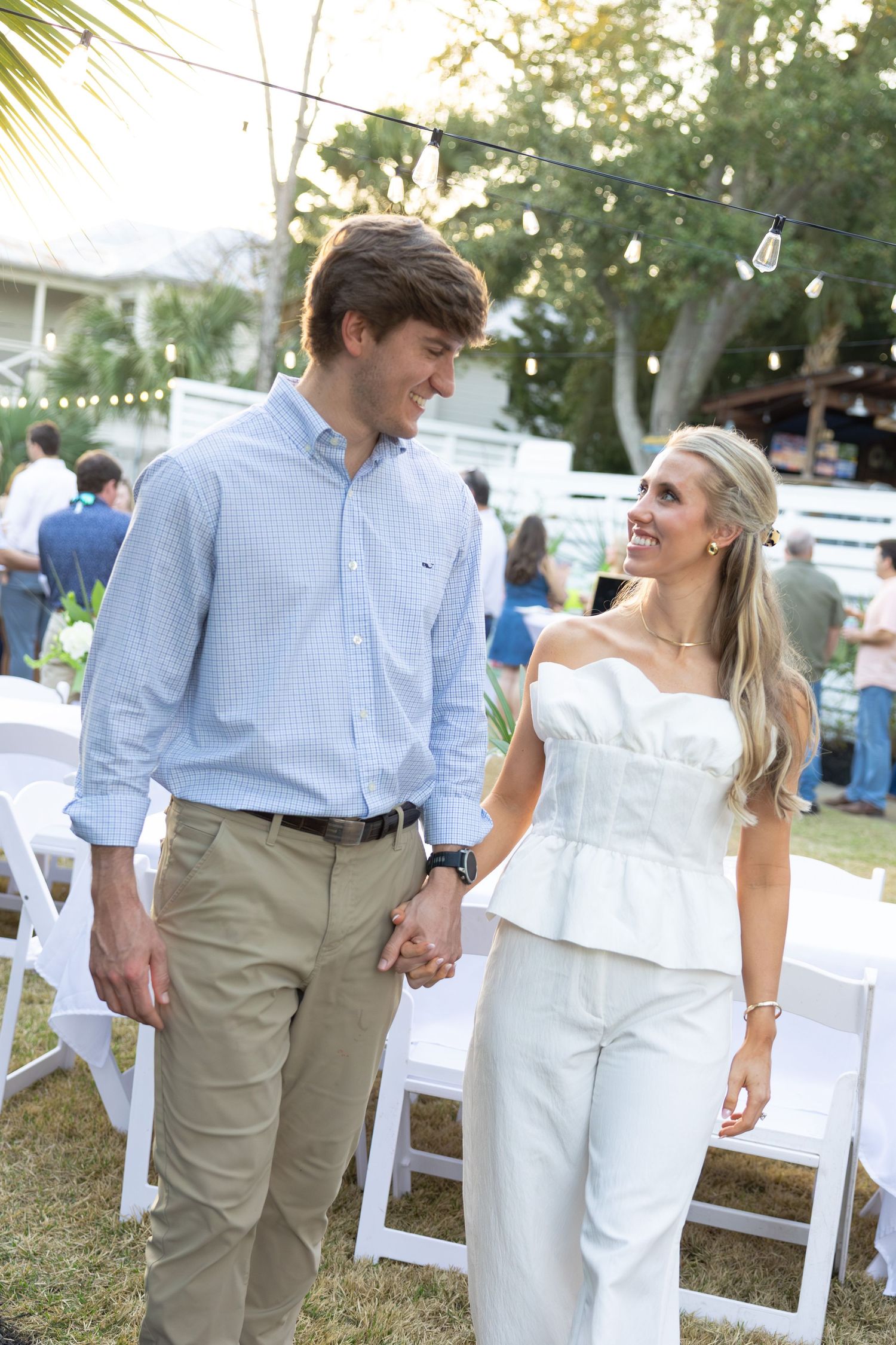 A couple in casual attire walks hand in hand through an outdoor event space with white chairs and palm trees.