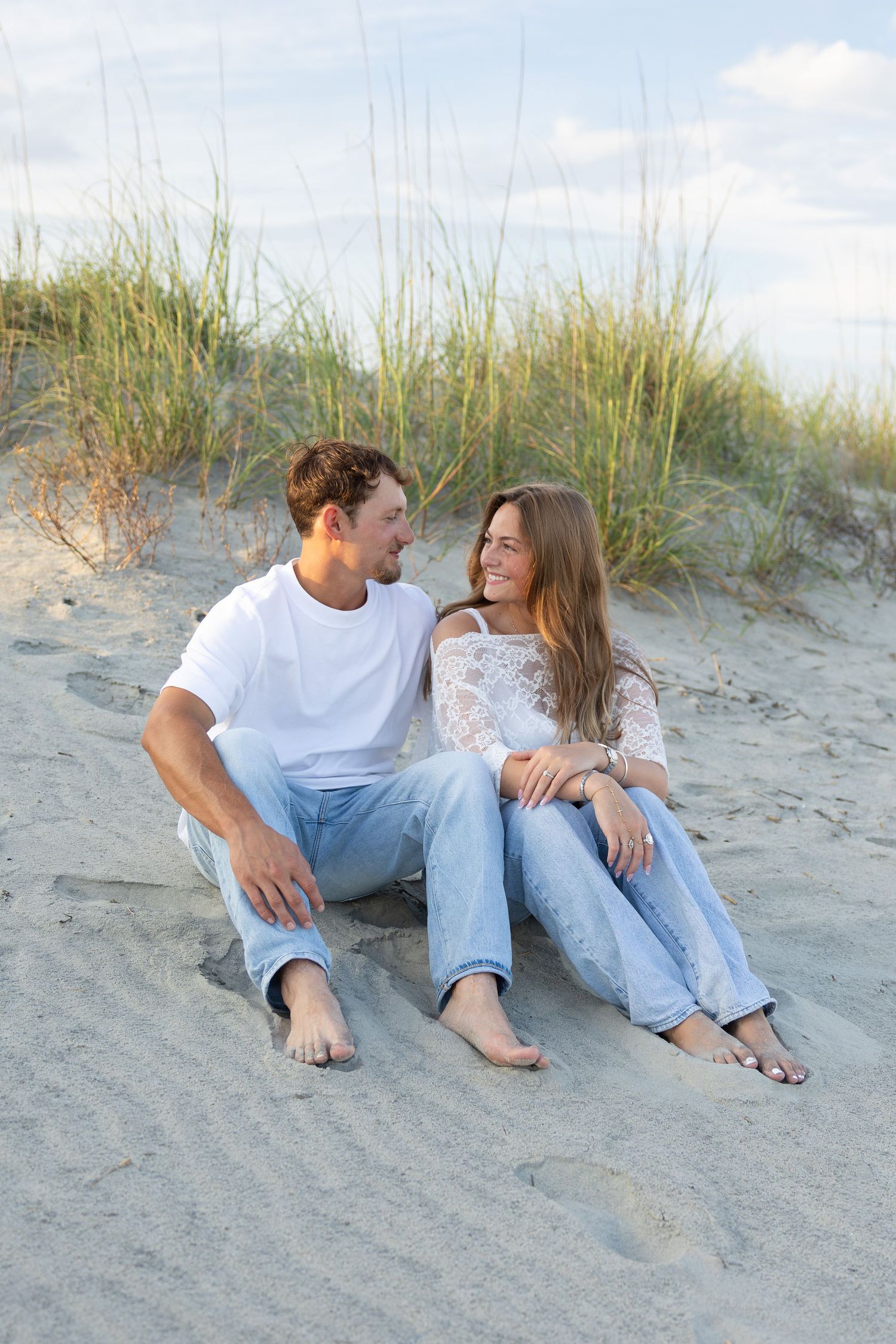 Happy couple sitting together on sandy beach dunes, wearing jeans and white tops, smiling at each other on a sunny day.