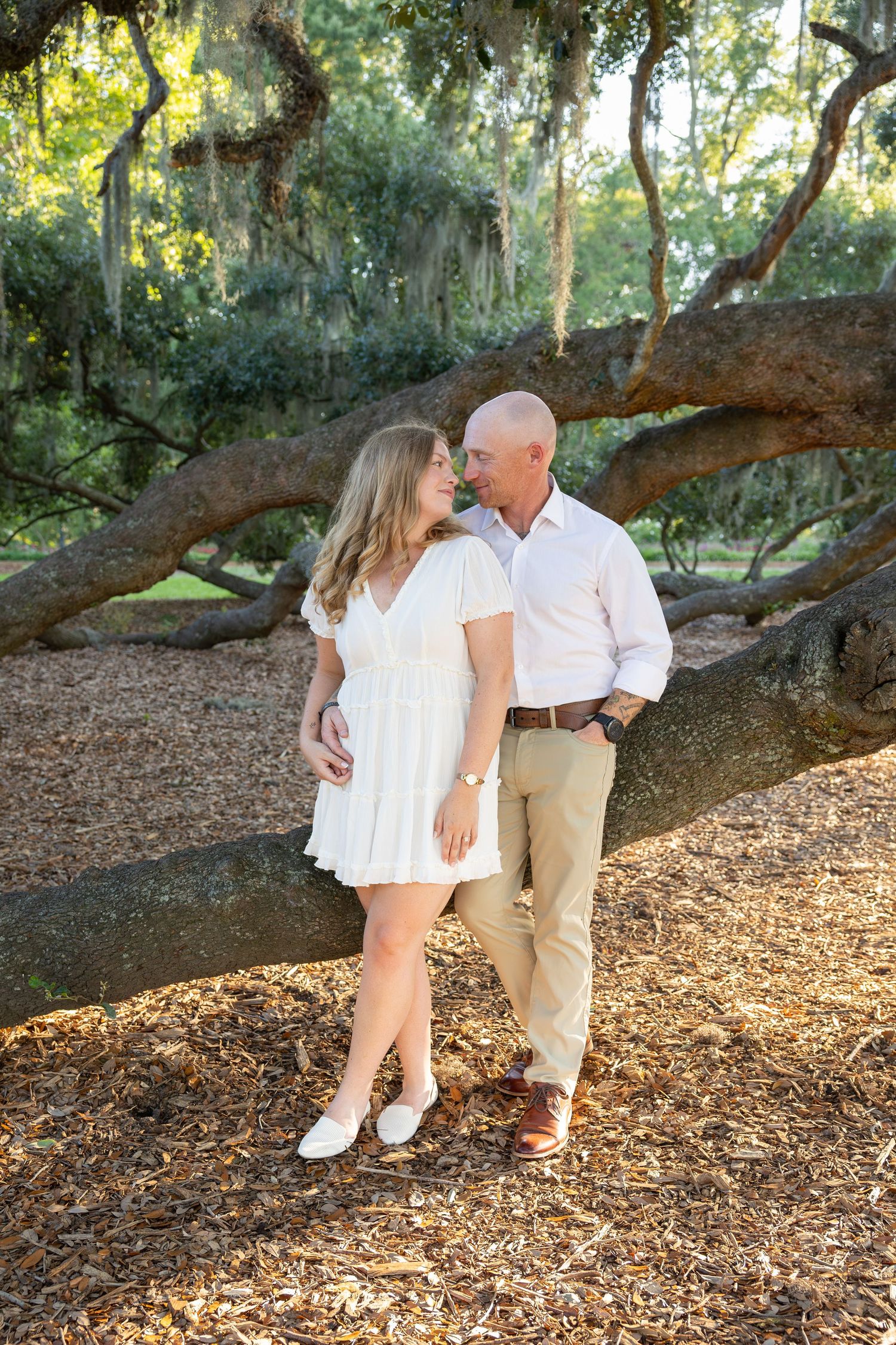 A couple shares a romantic moment under a sprawling oak tree with Spanish moss, woman in white dress, man in khakis.