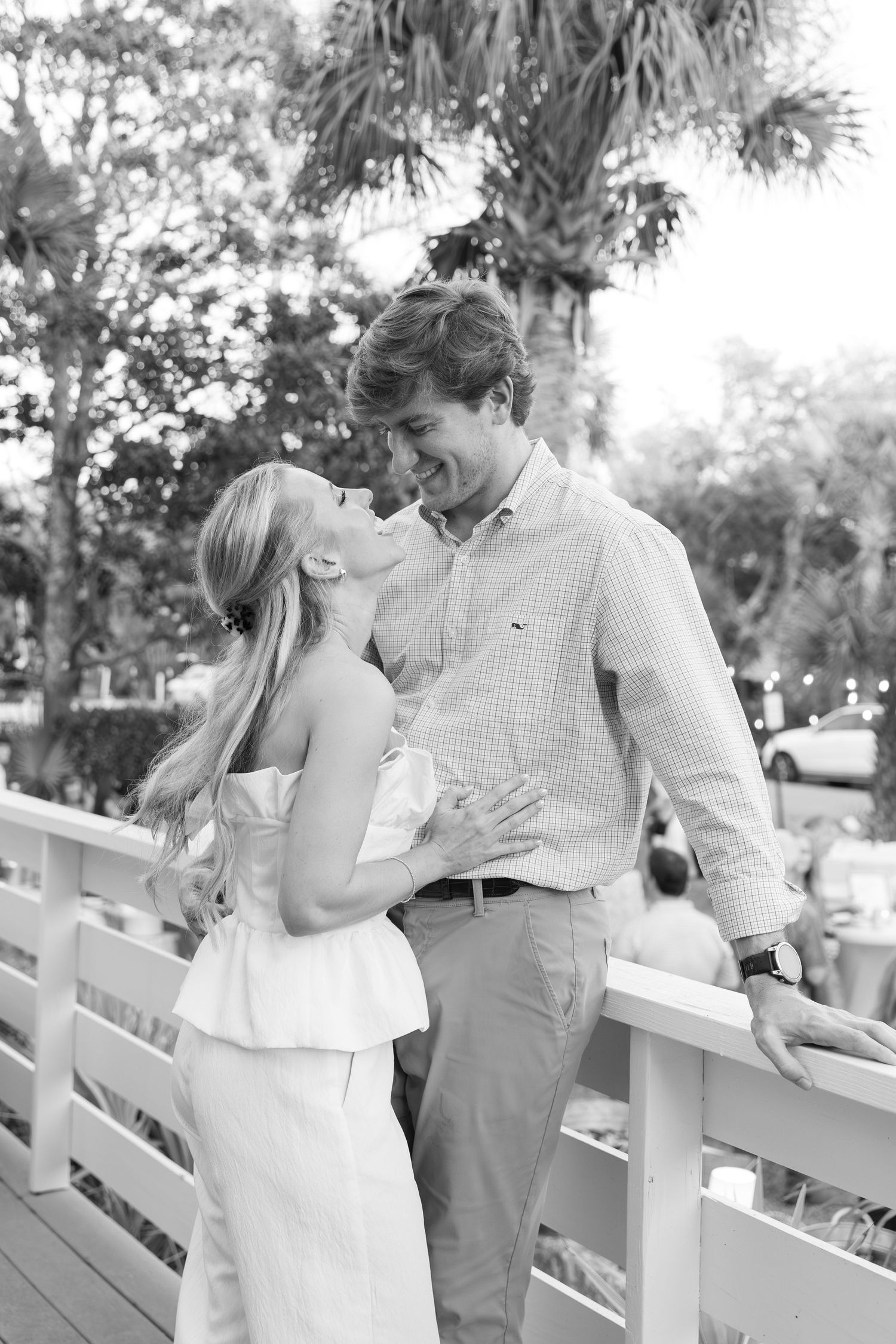 Black and white photo of a smiling couple sharing a romantic moment on a white railing bridge surrounded by trees.