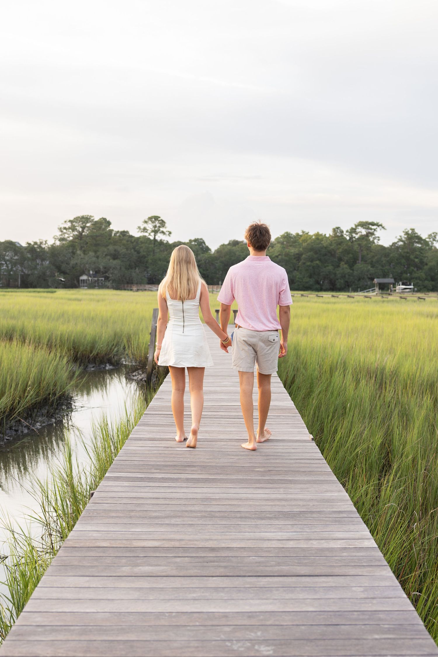 Couple holding hands walking barefoot on a wooden boardwalk through lush green marshlands.