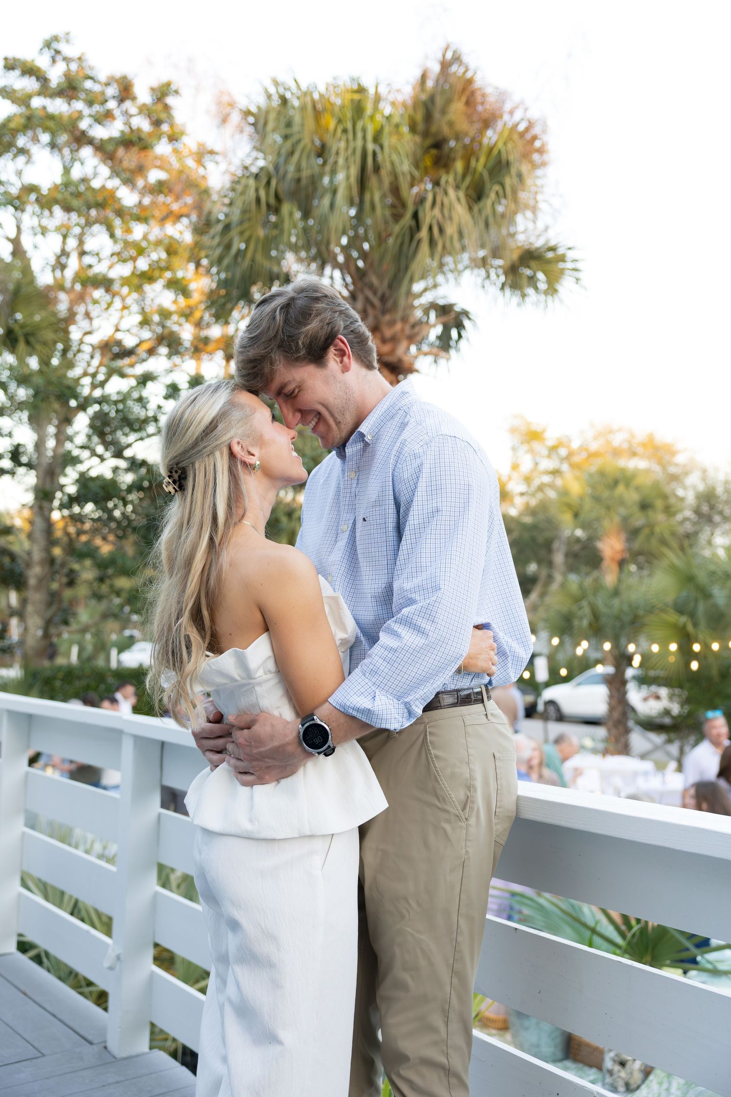 A romantic couple shares tender moments along a white fence during sunset as string lights glow in the background.