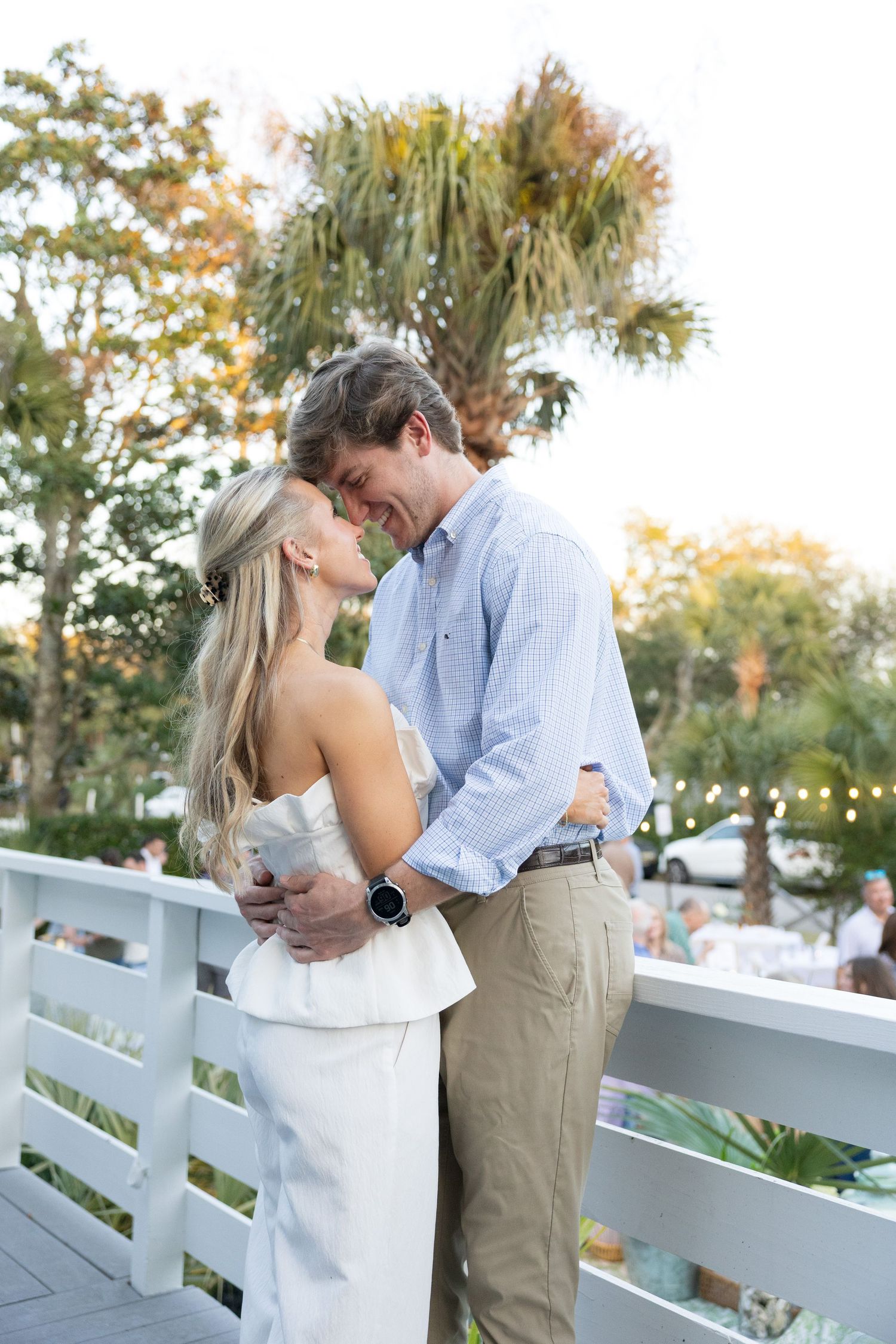 Couple sharing a romantic embrace on a white deck surrounded by palm trees at a coastal wedding venue.
